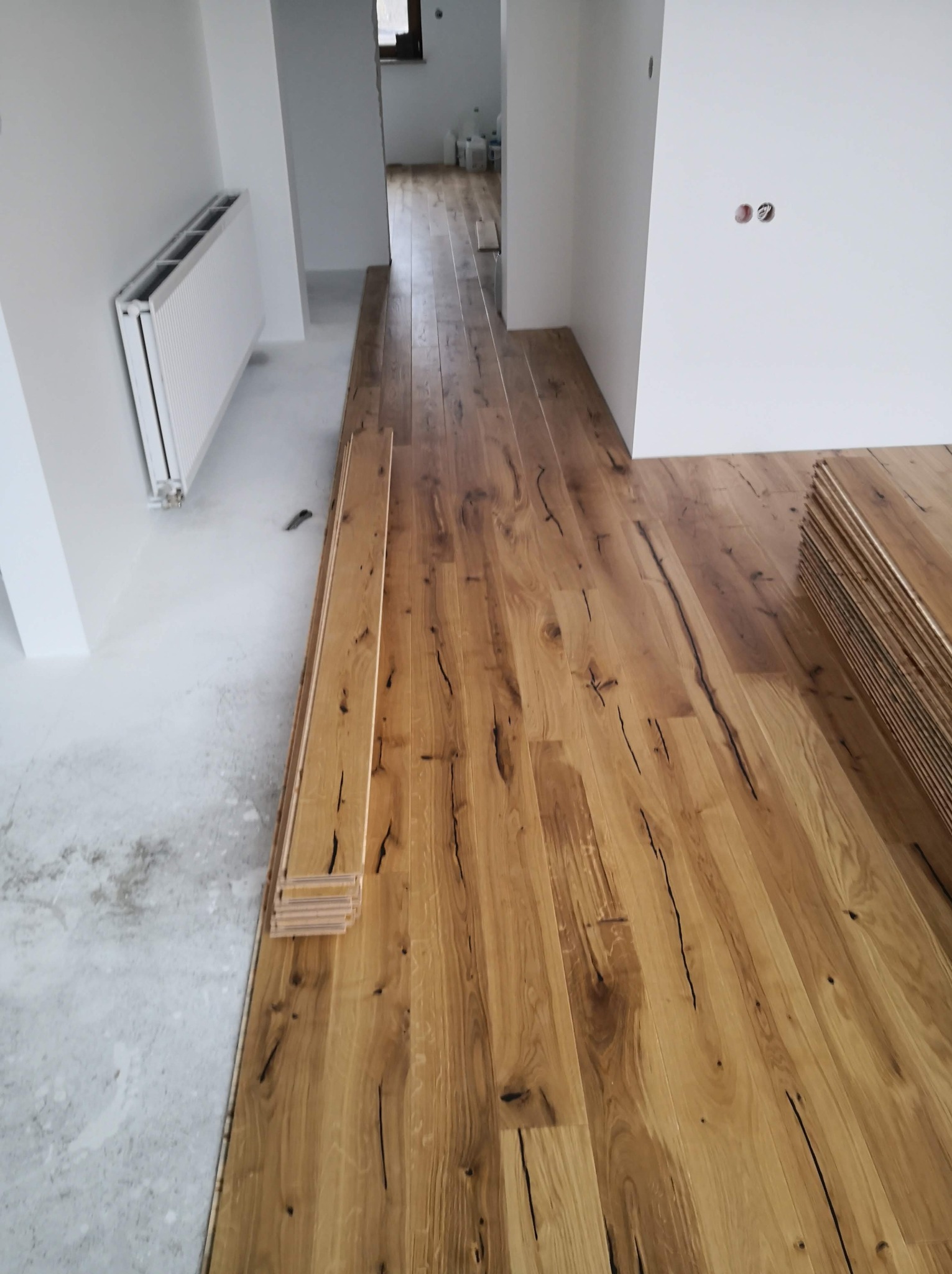 Układanie parkietu z wyraźnymi słojami i czarnymi pęknięciami w surowym, jasnym wnętrzu z widocznym grzejnikiem i przygotowanym podłożem.