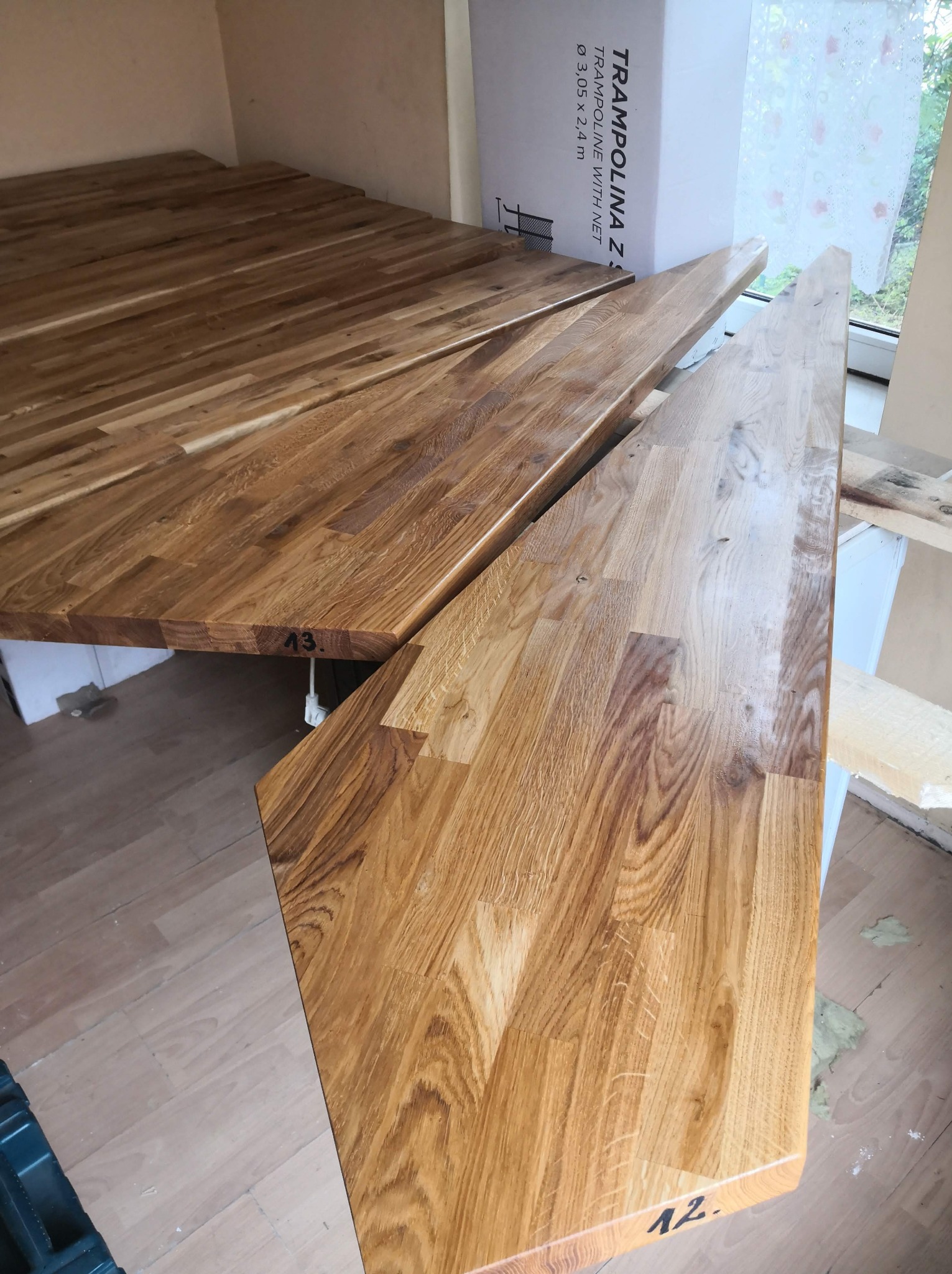 Fragmenty drewnianego parkietu lub paneli z widocznymi numerami '12' i '13' na krawędziach, ułożone na podłodze w pomieszczeniu w trakcie prac wykończeniowych, obok kartonu po trampolinie.