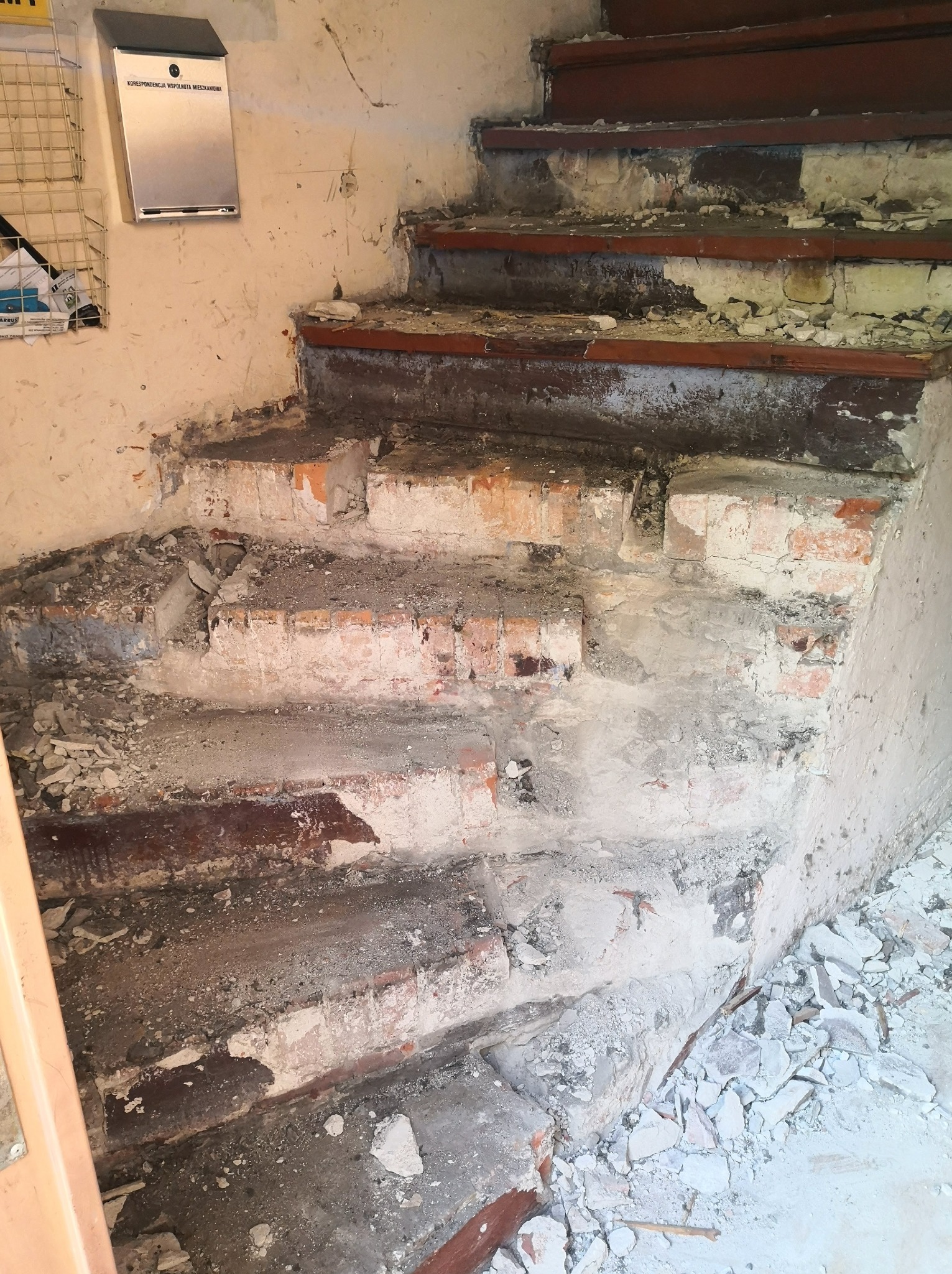 Schody w trakcie demontażu, widoczne cegły i resztki betonu, na ścianie skrzynka na listy.