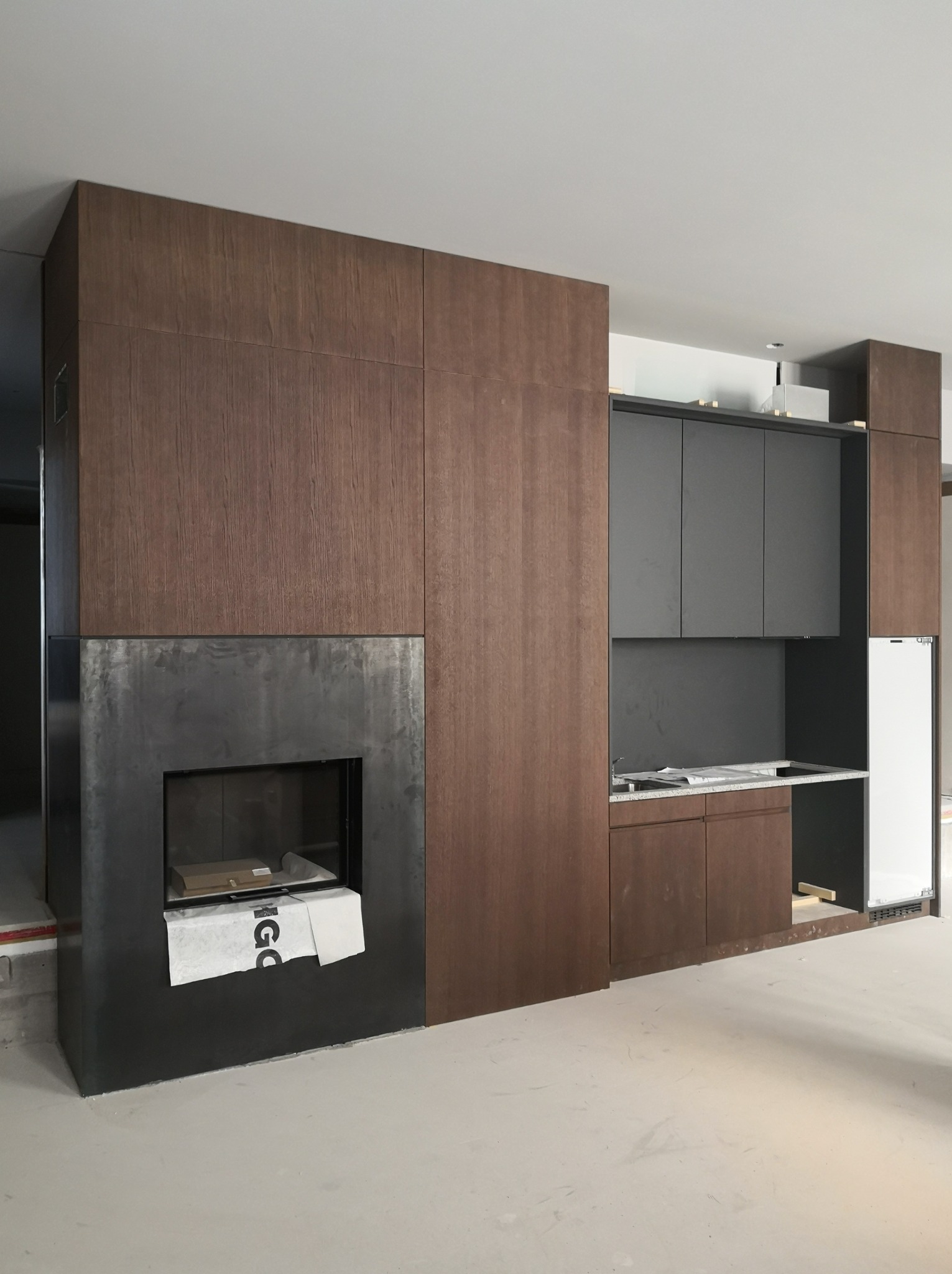 Wnętrze w trakcie prac wykończeniowych: zabudowa kominkowa z ciemnego drewna z metalowym wkładem, obok szare szafki kuchenne w zabudowie, na blacie leżą plany, podłoga wylewka.