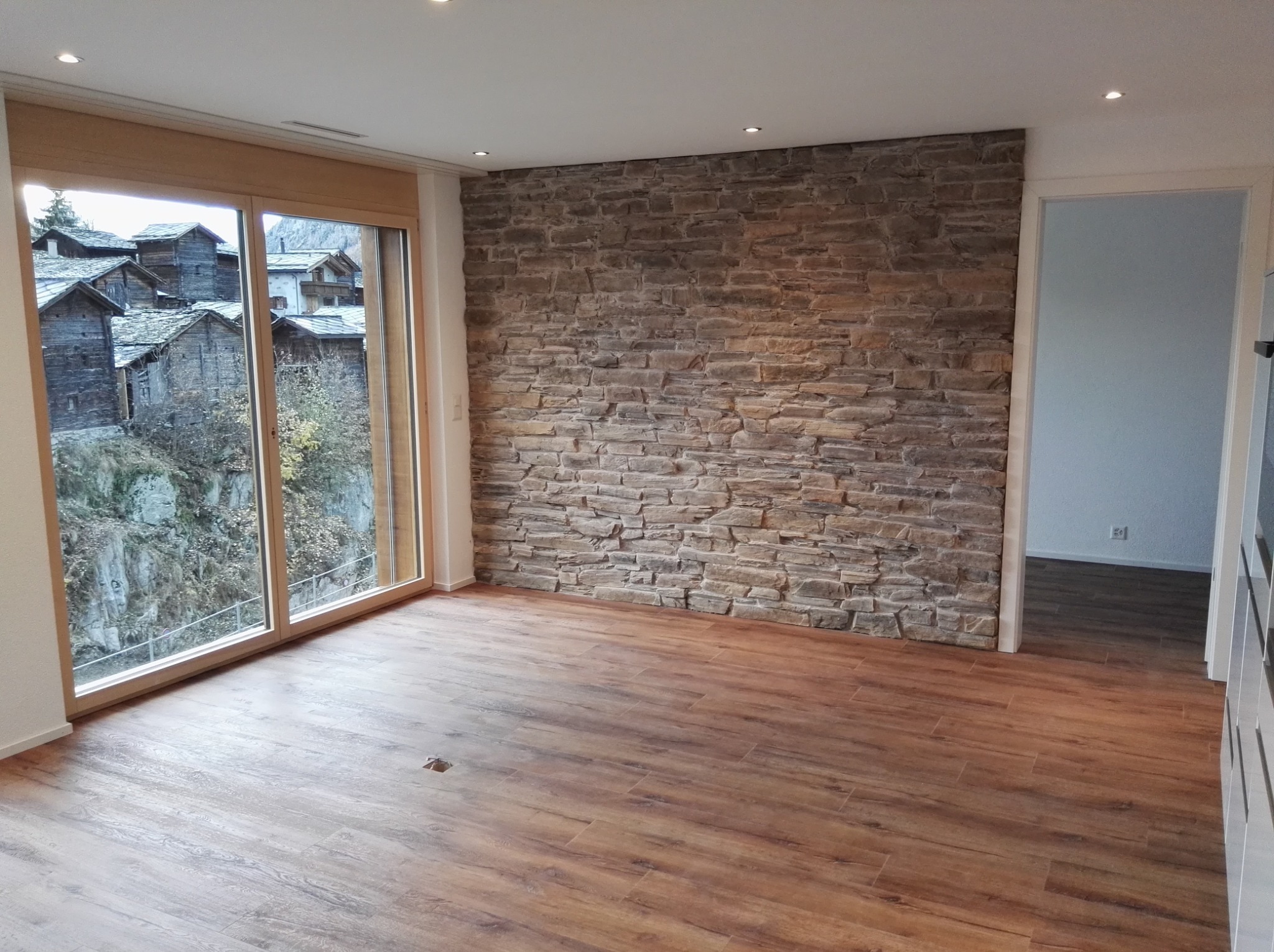 Wnętrze pokoju z drewnopodobną podłogą, kamienną ścianą i widokiem na górską wioskę przez duże okno.