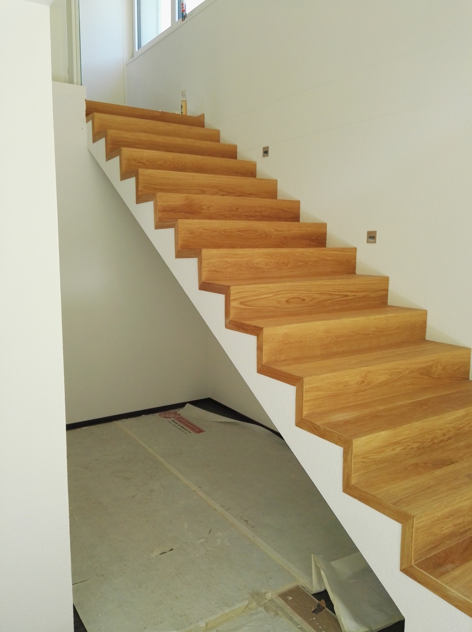 Drewniane schody z oświetleniem punktowym w ścianie, podłoga zabezpieczona folią ochronną, butelka na najwyższym stopniu.