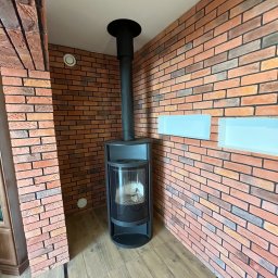 SKRZYD-BUD - Ceglana ściana z kominkiem w nowoczesnym wnętrzu. Widoczne płytki gipsowe imitujące cegłę, drewniana podłoga i minimalistyczne półki w ścianie.