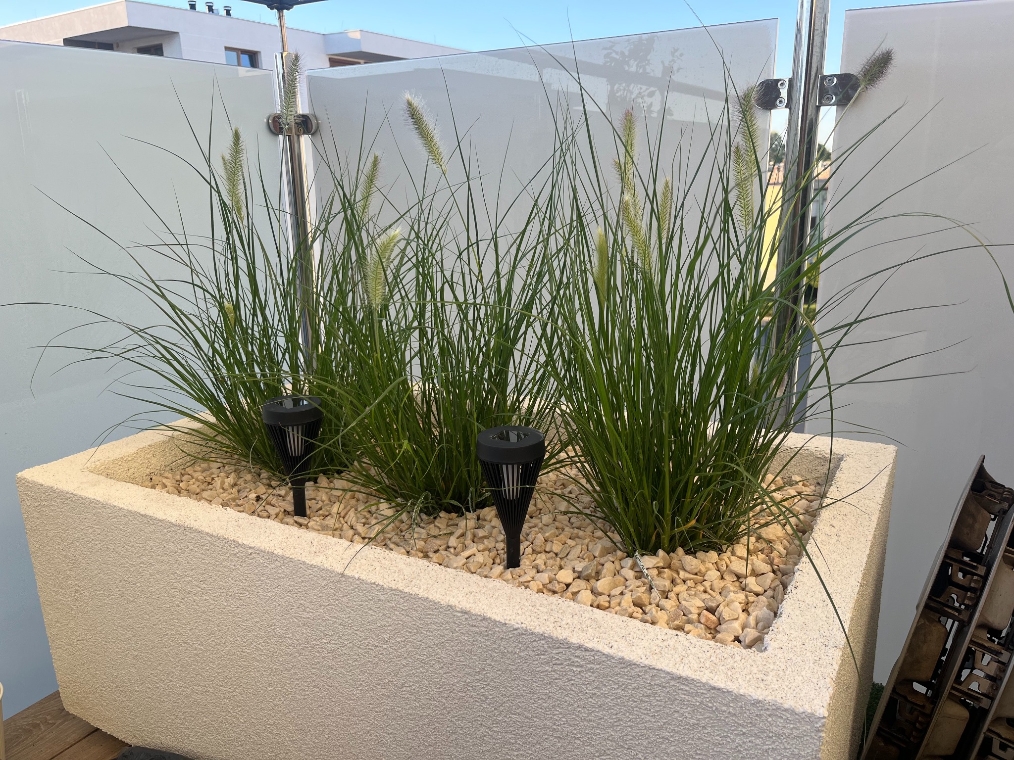 Donica z tynkiem silikonowym na balkonie, wypełniona kamieniami i trawą ozdobną, z lampkami solarnymi. Nowoczesne wykończenie balkonu z białymi panelami.