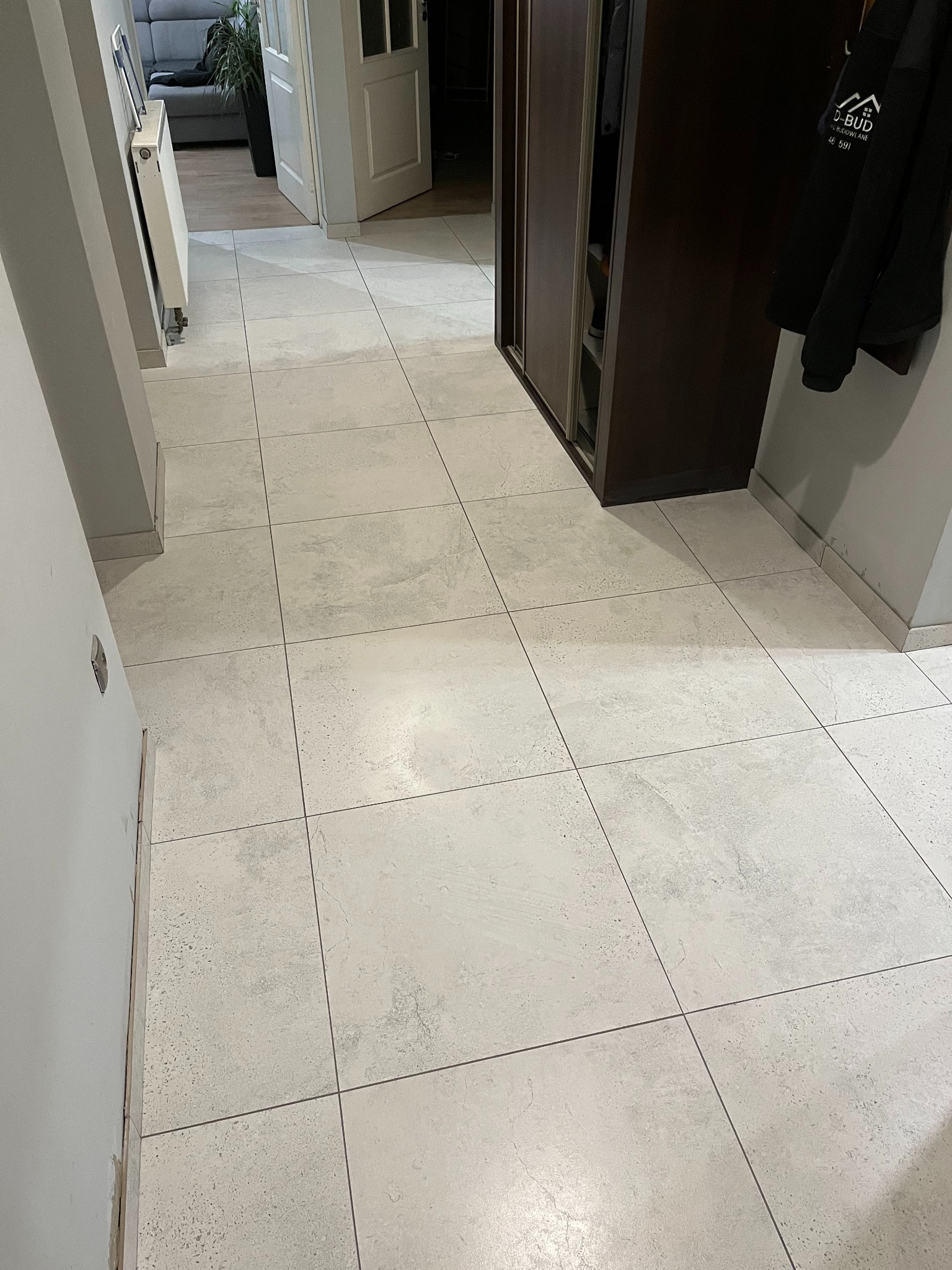 Jasne, matowe płytki podłogowe w korytarzu, ułożone równo z wąską fugą. W tle widoczna szafa przesuwna i fragment grzejnika.