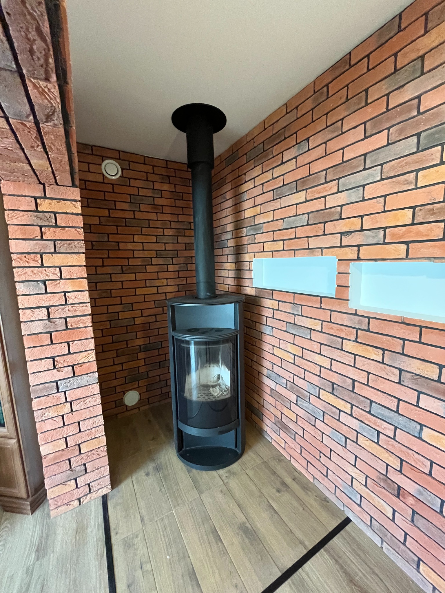 Ceglana ściana z kominkiem w nowoczesnym wnętrzu. Widoczne płytki gipsowe imitujące cegłę, drewniana podłoga i minimalistyczne półki w ścianie.