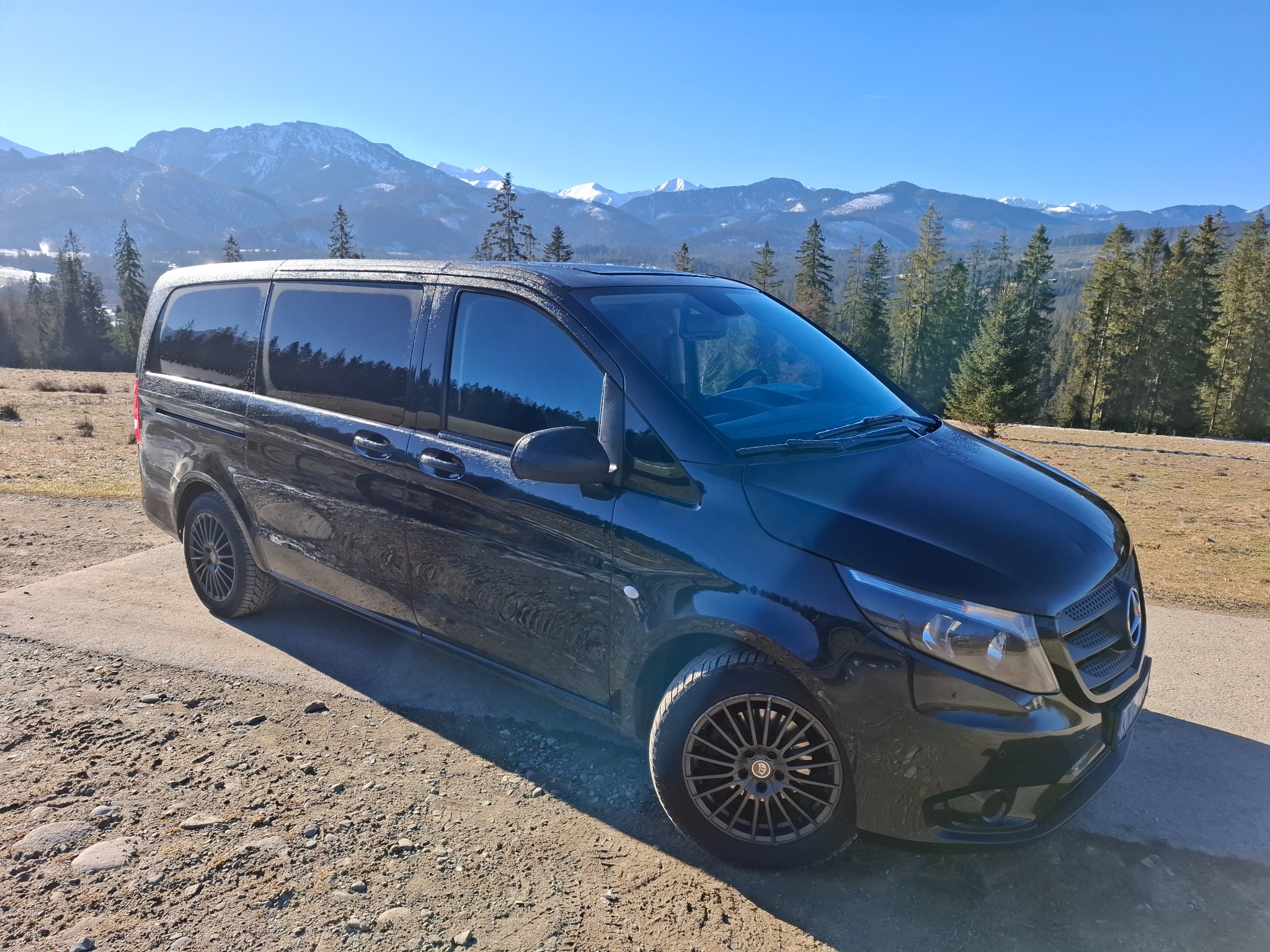 Czarny van Mercedes-Benz Vito z przyciemnianymi szybami na tle górskiego krajobrazu z ośnieżonymi szczytami i lasem iglastym w słoneczny dzień.