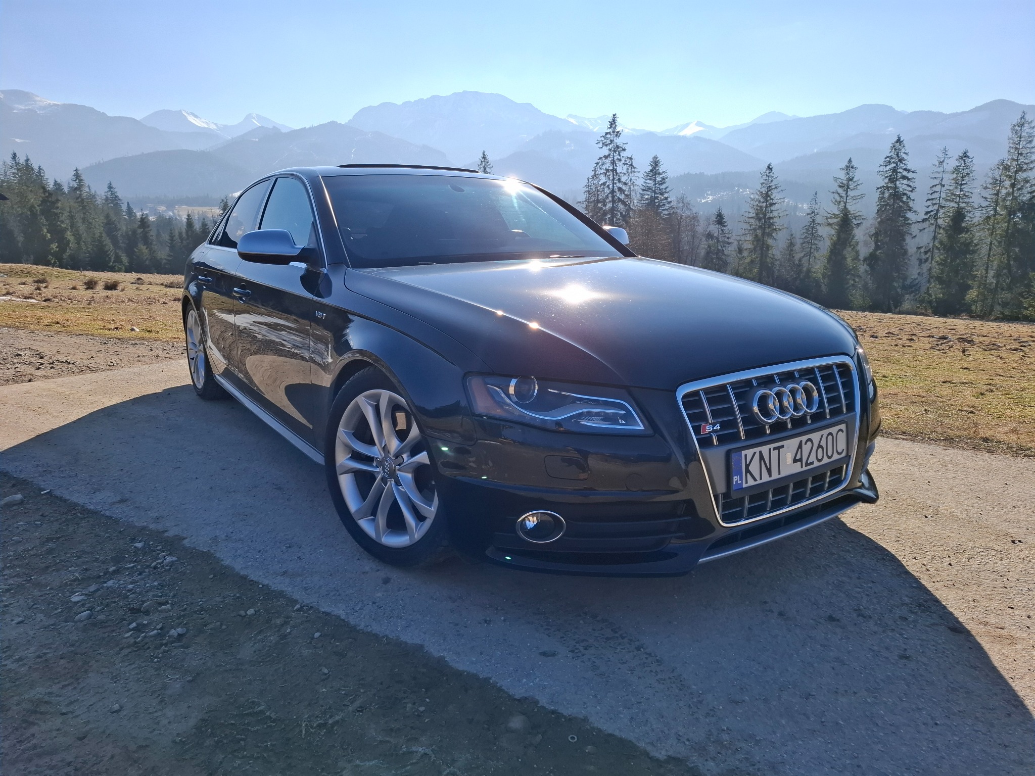Czarny samochód Audi S4 z polską tablicą rejestracyjną KNT 4260C zaparkowany na szutrowej drodze z widokiem na zalesione wzgórza i ośnieżone szczyty górskie w tle, w słoneczny dzień.