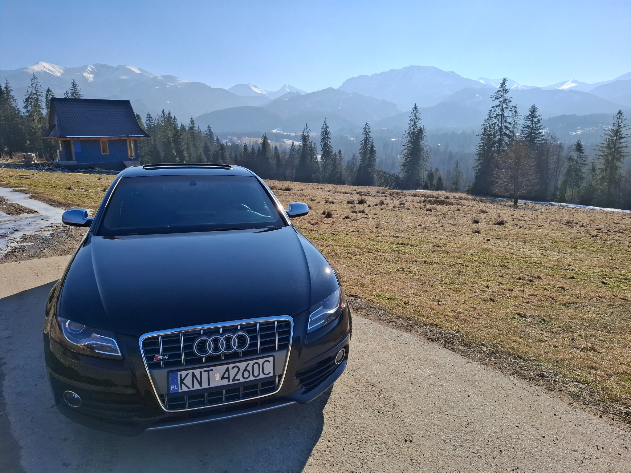 Czarny samochód Audi S4 z polską tablicą rejestracyjną KNT 4260C na tle malowniczego krajobrazu górskiego z domkiem z niebieskimi ścianami.