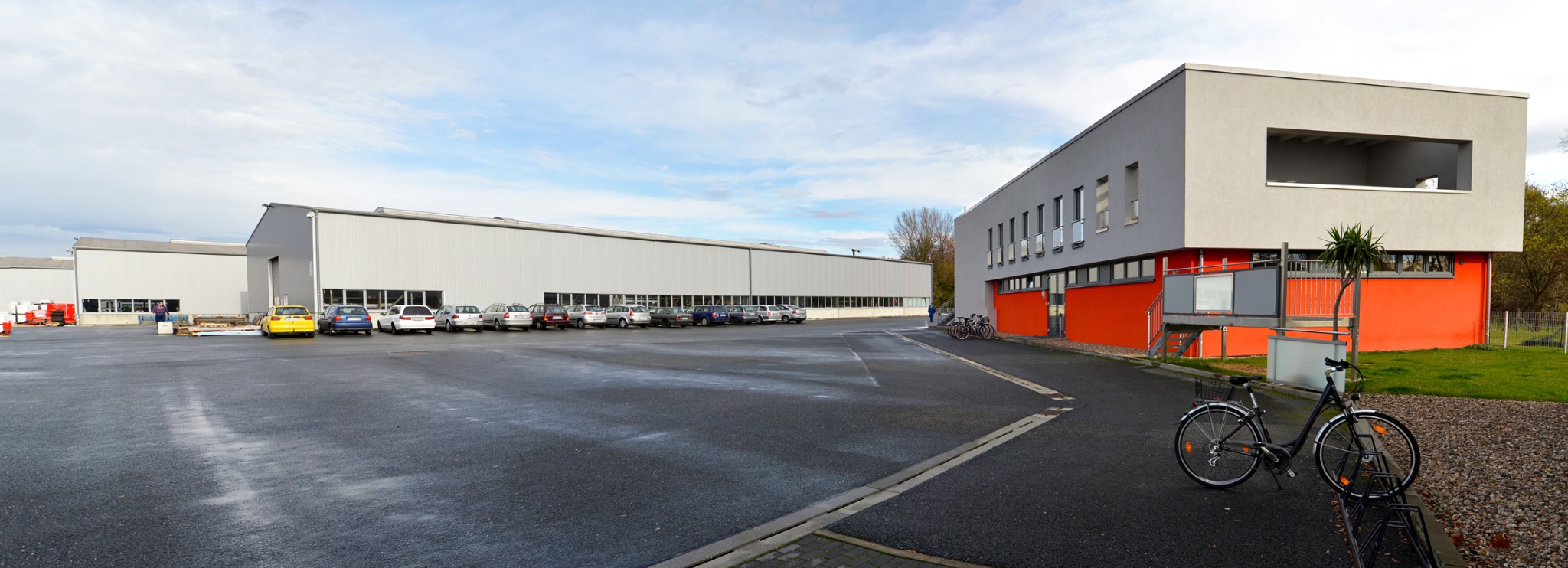 Szeroki widok zewnętrzny nowoczesnego budynku przemysłowego z szarymi ścianami i pomarańczowym akcentem, z rzędem zaparkowanych samochodów i rowerami w stojaku z przodu.