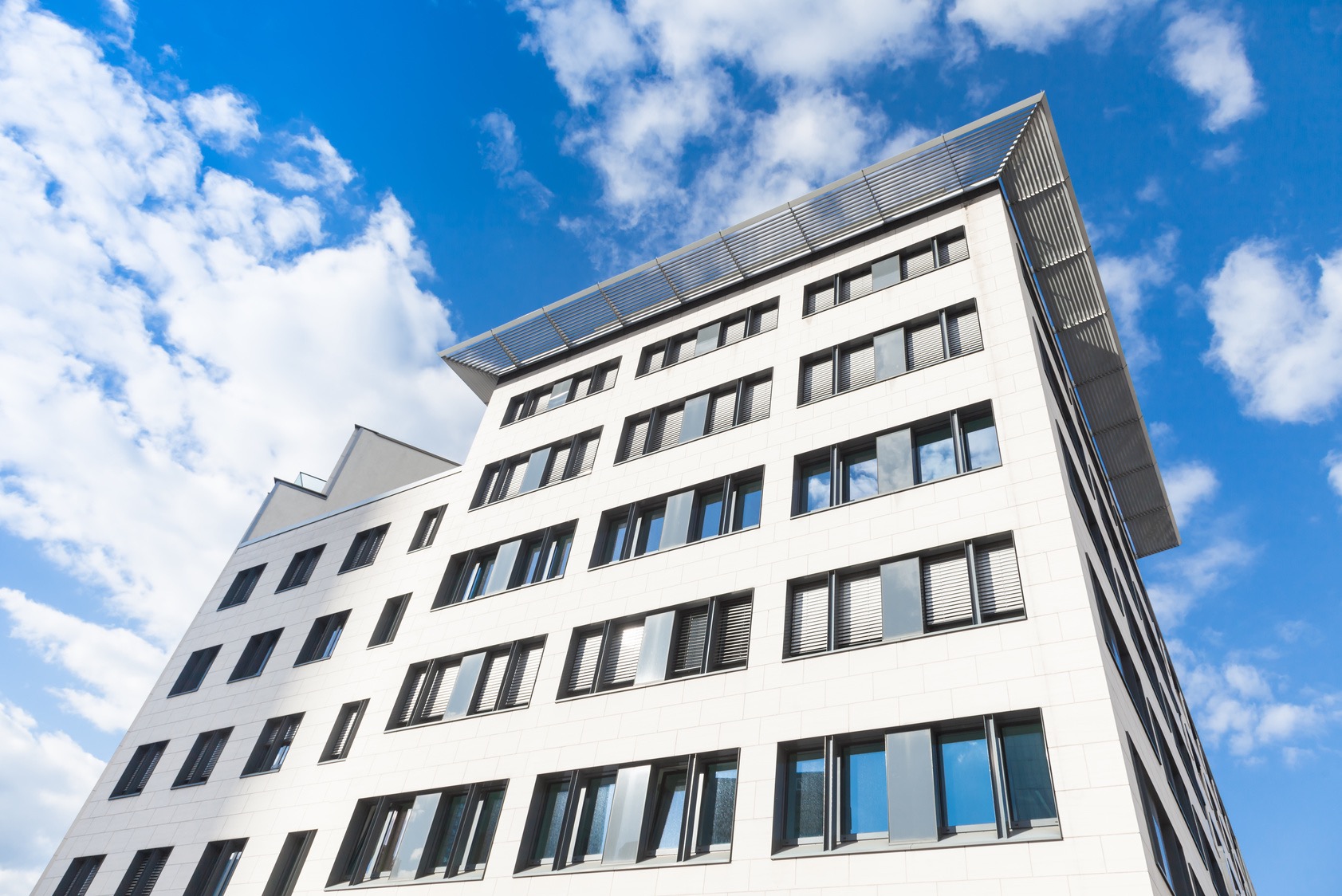 Nowoczesny budynek biurowy z białej cegły, z metalowym zadaszeniem, widziany z dołu na tle błękitnego nieba z chmurami.