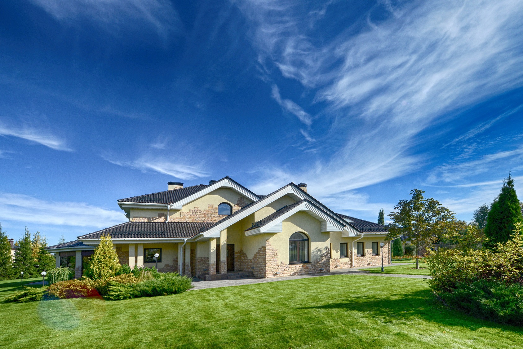 Nowoczesny, dwukondygnacyjny dom z beżową elewacją i elementami z kamienia, otoczony zadbanym trawnikiem i dekoracyjnymi krzewami, na tle błękitnego nieba z cirrusami.