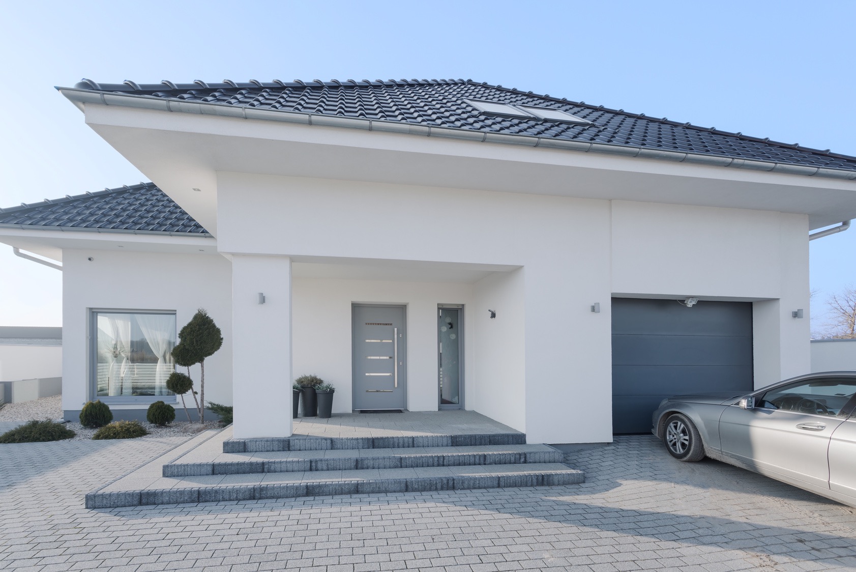 Nowoczesny, biały dom jednorodzinny z szarą stolarką okienną i drzwiową, oraz z garażem z bramą segmentową, widok z zewnątrz.