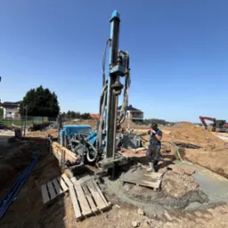 Niebieska wiertnica na placu budowy, pracownik w kombinezonie obsługuje maszynę, w tle widoczny wykop i koparka.