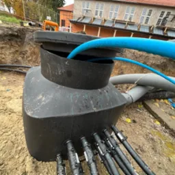 Podziemny zbiornik z podłączonymi rurami w wykopie w Tarnowcu, widoczne oznaczenia na rurach i zbiorniku, budynek w tle.