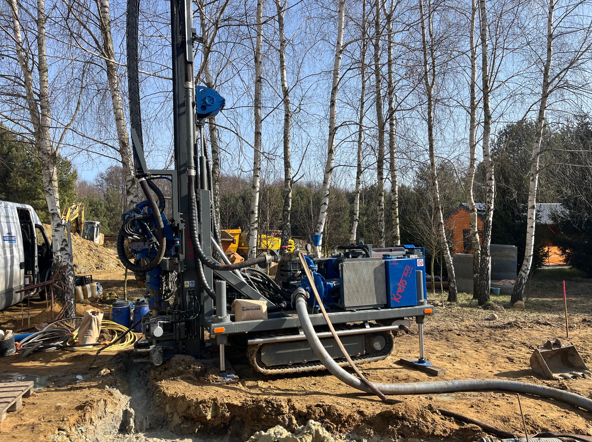 Wiertnica geologiczna KRETO MAX na gąsienicach, pracująca na placu budowy, z widocznymi brzozami w tle i wykopem z wodą.