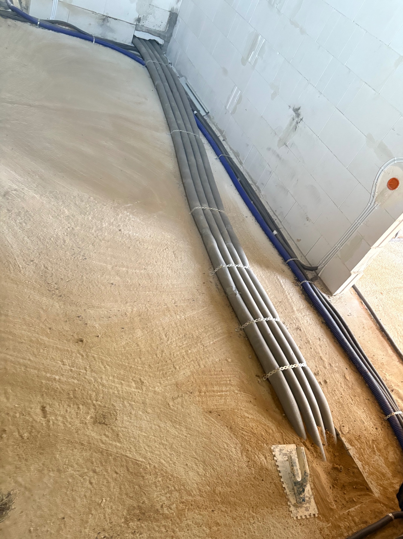 Rury pompy ciepła w izolacji termicznej, prowadzone w bruzdach w ścianie i podłodze, oraz niebieskie rury karbowane, widoczna paca do gładzi.
