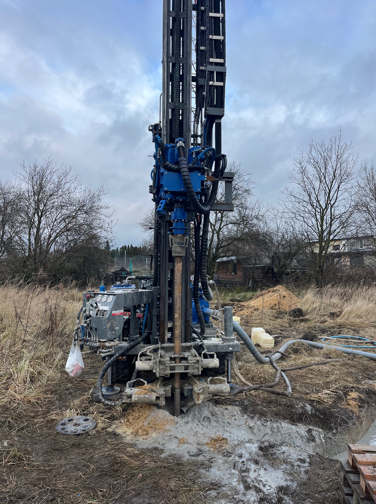 Wiertnica do odwiertów pionowych w trakcie pracy na zewnątrz, widoczne błoto i osprzęt, pochmurne niebo w tle.