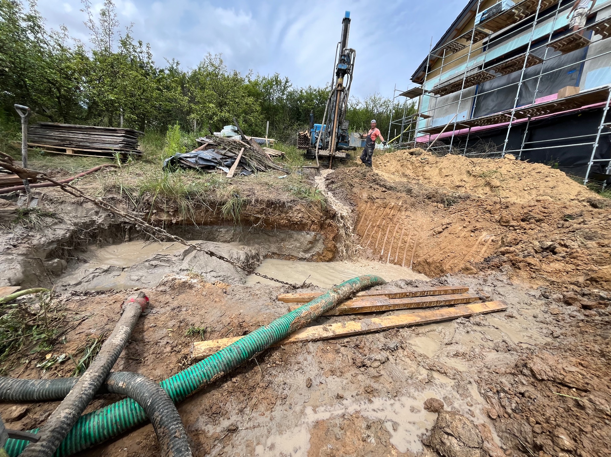 Wiertnica do studni w trakcie pracy na budowie domu jednorodzinnego, widoczny wykop wypełniony wodą, zielone węże, pracownik w pomarańczowej koszulce oraz rusztowanie przy budynku.