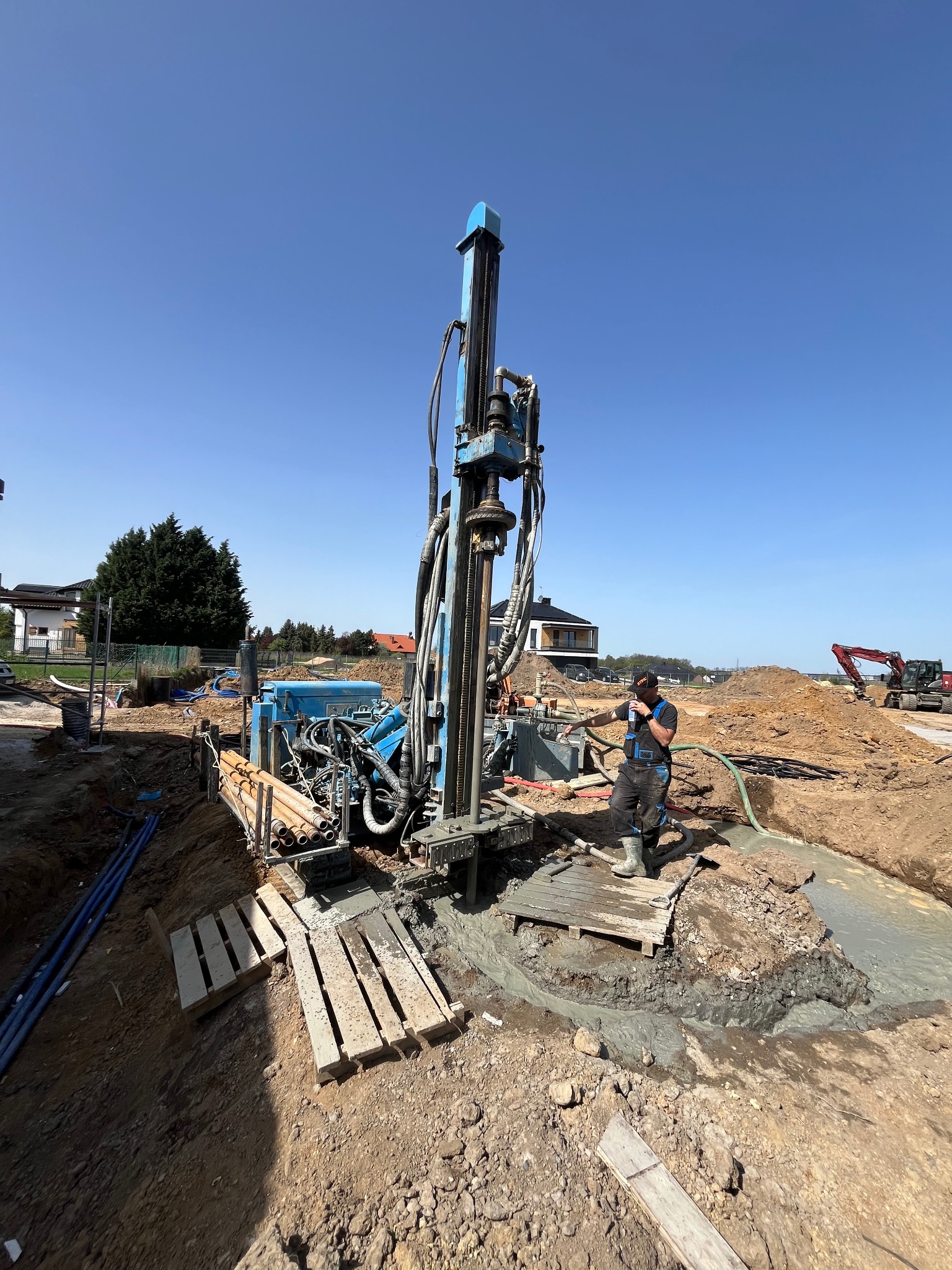 Niebieska wiertnica na placu budowy, pracownik w kombinezonie obsługuje maszynę, w tle widoczny wykop i koparka.