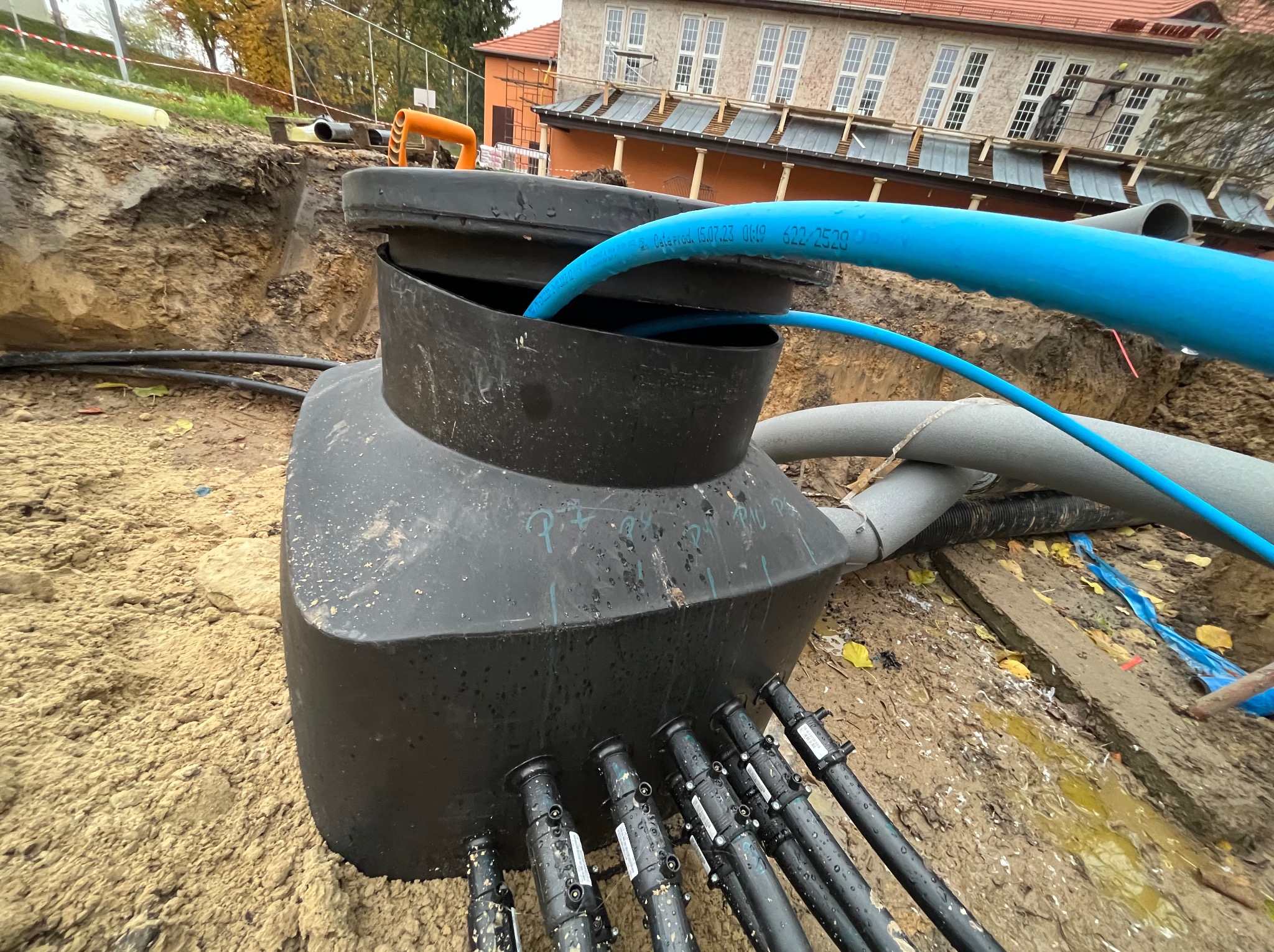 Podziemny zbiornik z podłączonymi rurami w wykopie w Tarnowcu, widoczne oznaczenia na rurach i zbiorniku, budynek w tle.