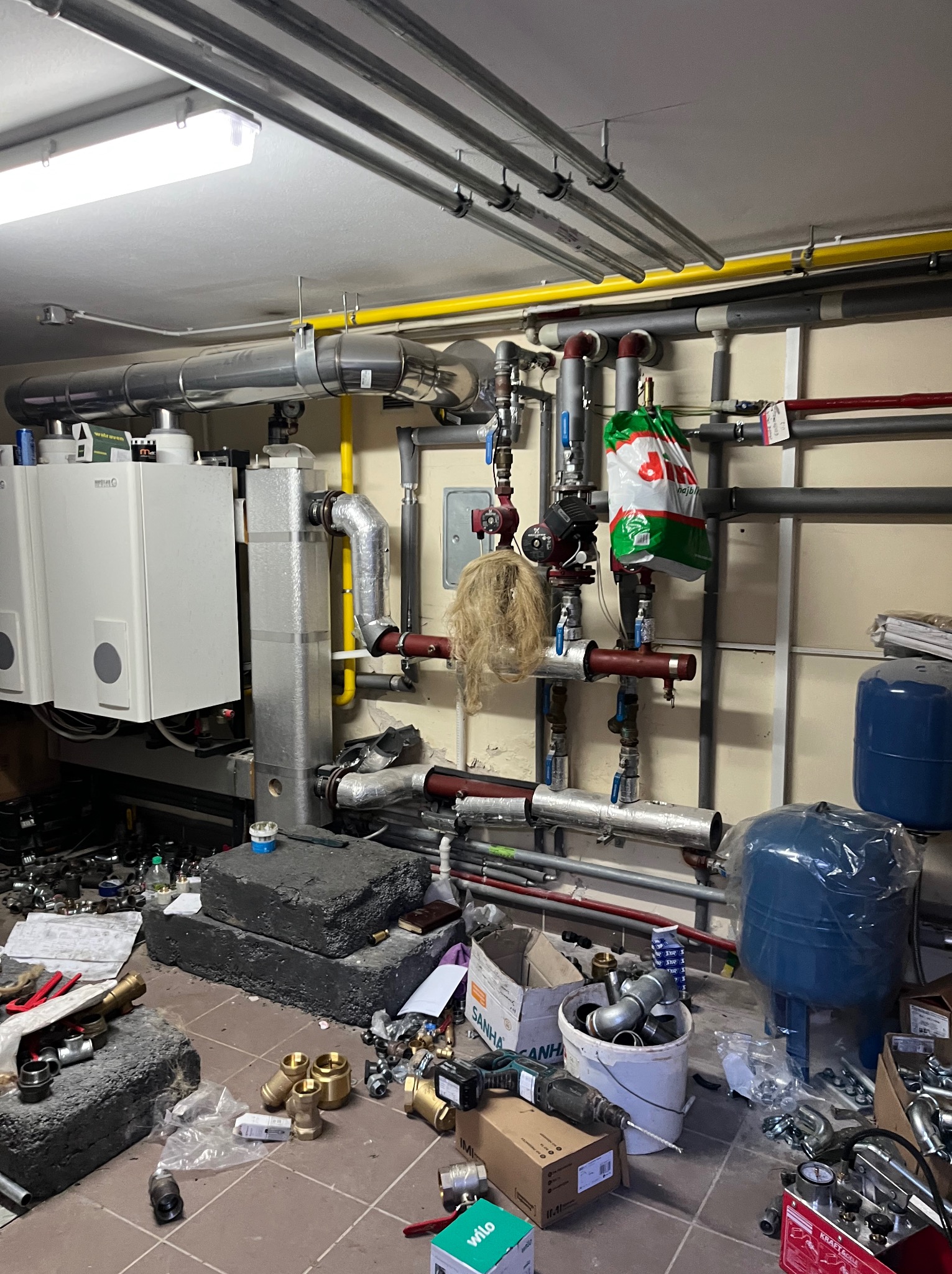 Kotłownia w trakcie montażu: dwa białe kotły grzewcze, instalacja rur, armatura, zbiorniki wyrównawcze i narzędzia, widoczne elementy instalacji centralnego ogrzewania i wodno-kanalizacyjnej.