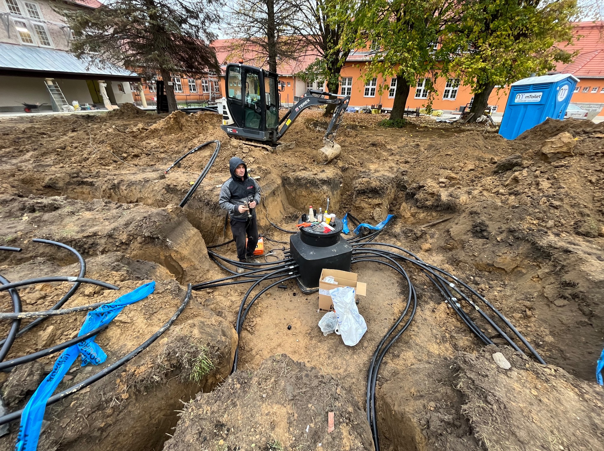 Instalacja podziemna z czarną skrzynką rozdzielczą i licznymi kablami w wykopie ziemnym, z operatorem minikoparki w tle na tle budynku.