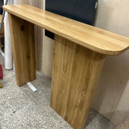 BAYWOOD - Konstrukcja z jasnego drewna o zaokrąglonych krawędziach, przypominająca konsolę lub stolik pomocniczy, widoczna na tle warsztatu stolarskiego z elementami wyposażenia.