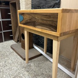 BAYWOOD - Stolik nocny z drewna jesionowego z szufladą z orzecha włoskiego i wstawką z żywicy epoksydowej, na tle warsztatu stolarskiego z widoczną komodą i drapakiem dla kota.