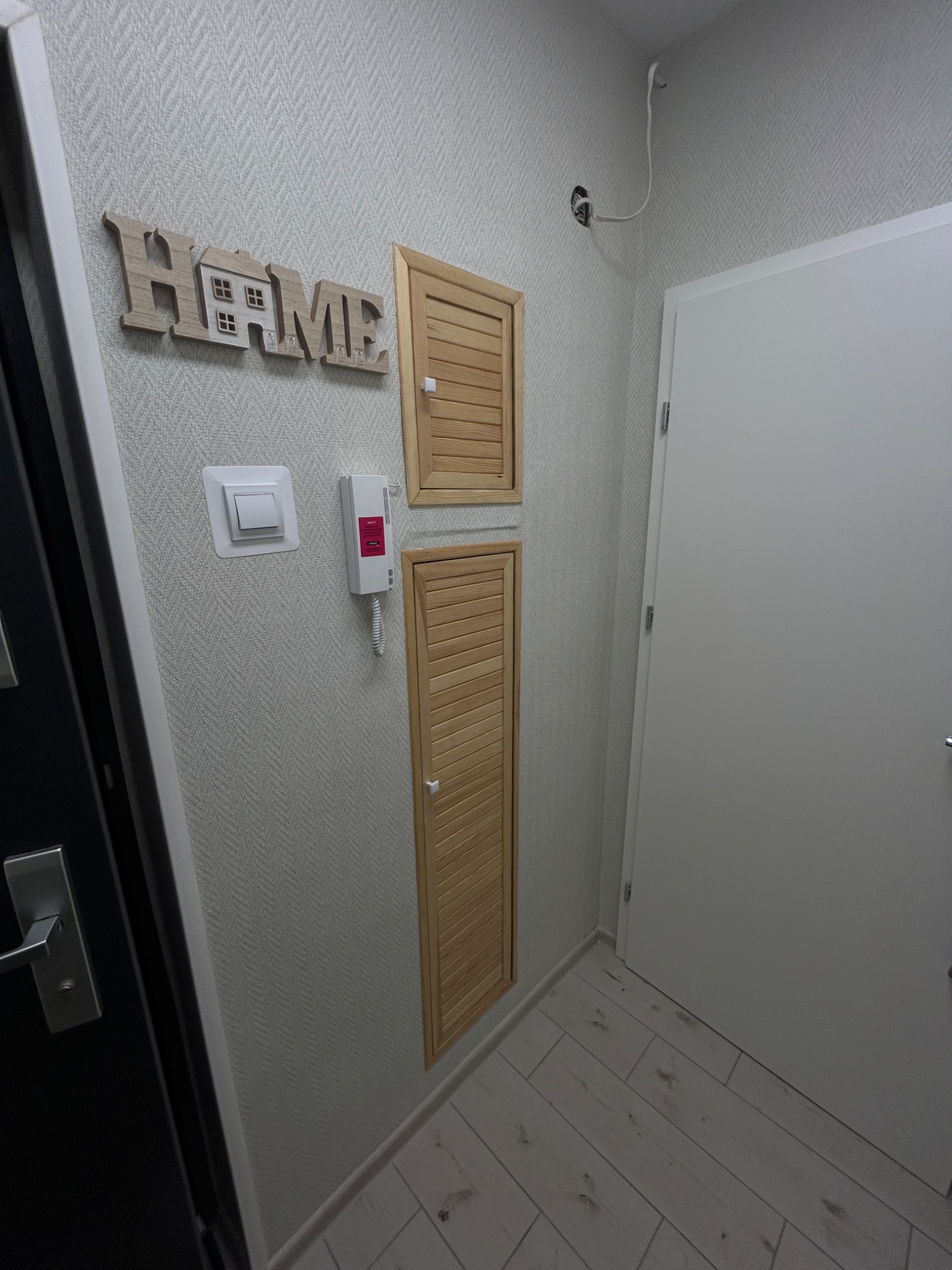 Korytarz z drewnianymi drzwiczkami rewizyjnymi do instalacji, białą ścianą z dekoracyjną tapetą, drewnianym napisem HOME i białą szafą wnękową.