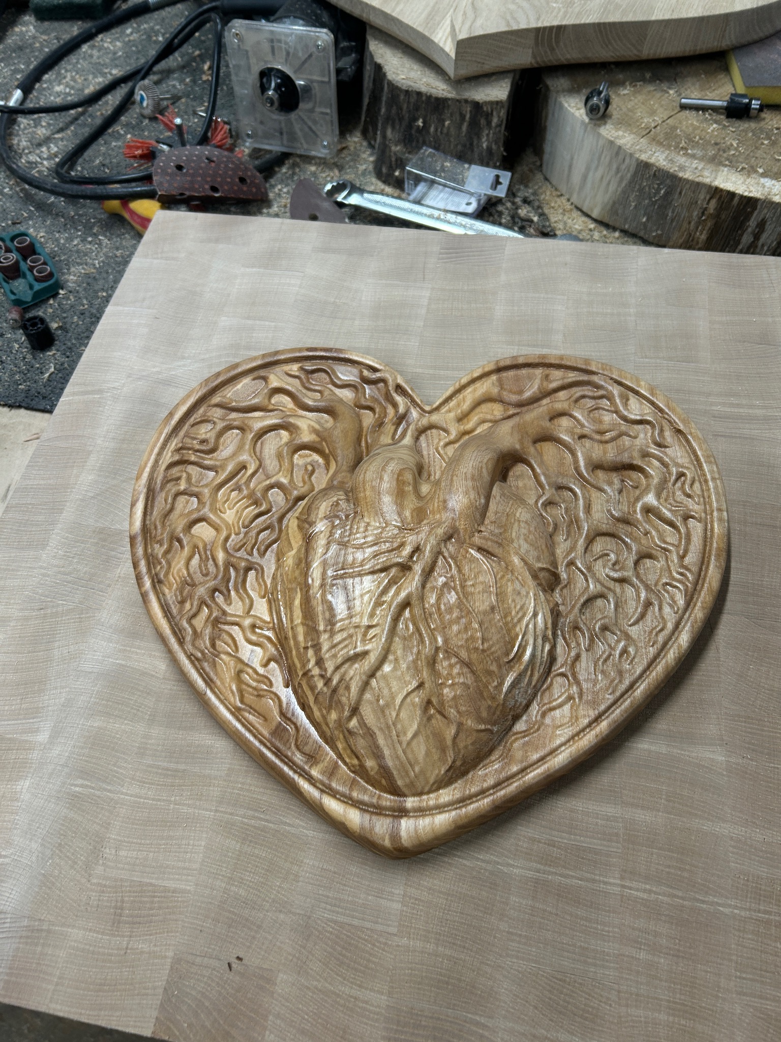 Ręcznie rzeźbiona drewniana płaskorzeźba w kształcie serca z motywem anatomicznego serca i korzeni drzewa, leżąca na drewnianym blacie w warsztacie.