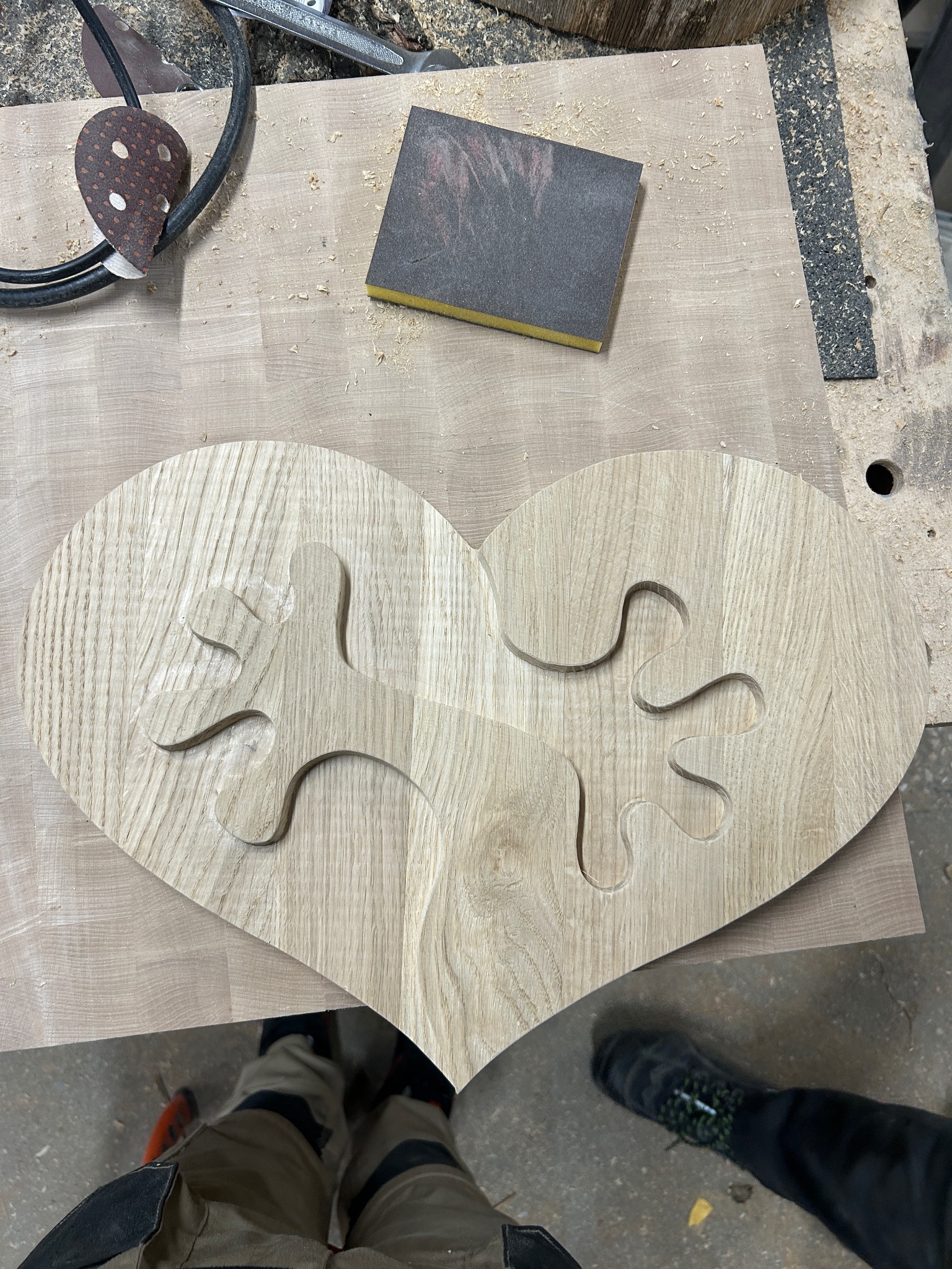 Drewniane serce z wyciętymi puzzlami, leżące na warsztacie, obok narzędzi do obróbki drewna, w tle widoczne nogi osoby fotografującej.