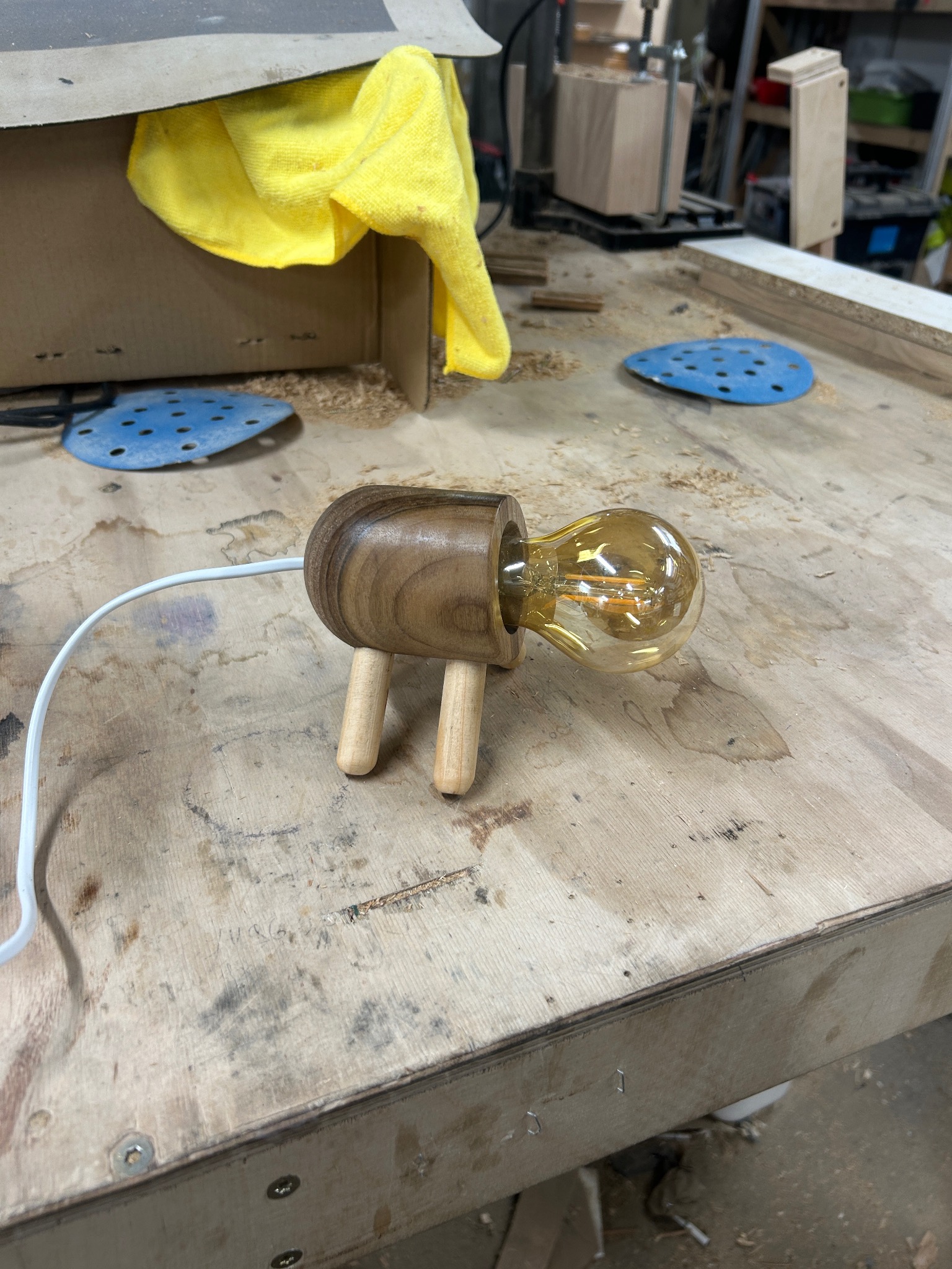 Drewniana lampka nocna w kształcie zwierzątka z żarówką Edison na warsztacie w Katowicach, w tle narzędzia i materiały stolarskie.