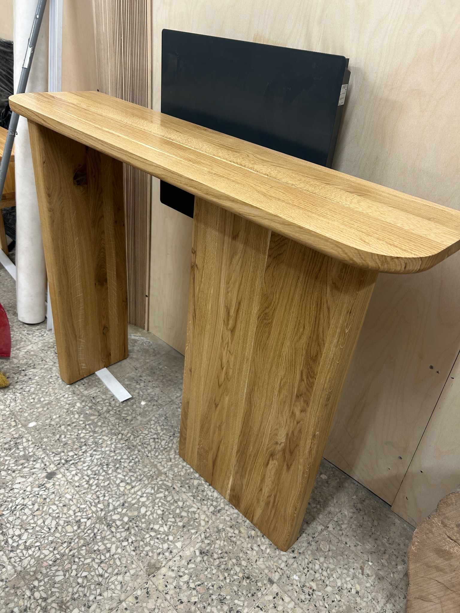 Konstrukcja z jasnego drewna o zaokrąglonych krawędziach, przypominająca konsolę lub stolik pomocniczy, widoczna na tle warsztatu stolarskiego z elementami wyposażenia.