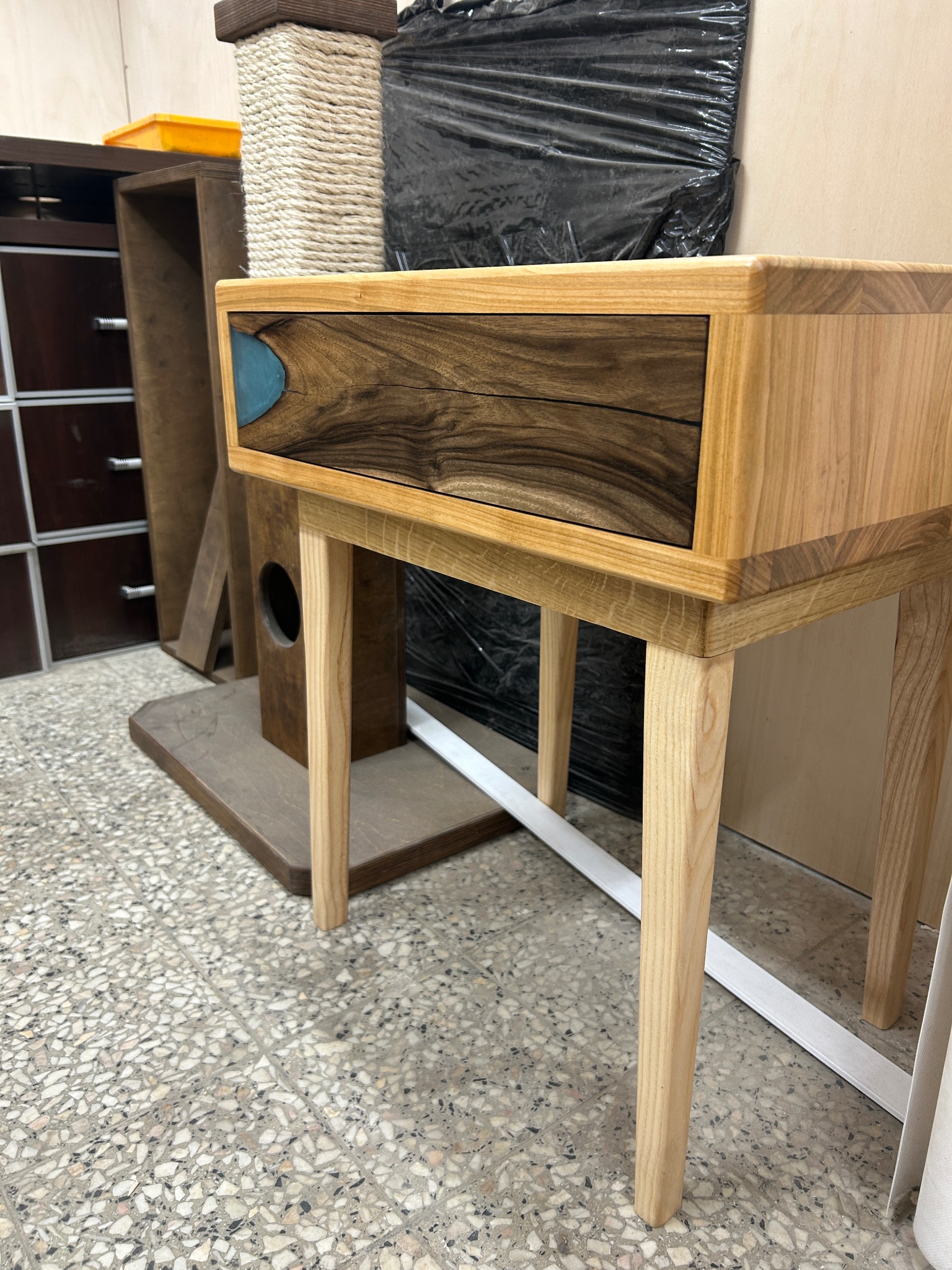 Stolik nocny z drewna jesionowego z szufladą z orzecha włoskiego i wstawką z żywicy epoksydowej, na tle warsztatu stolarskiego z widoczną komodą i drapakiem dla kota.
