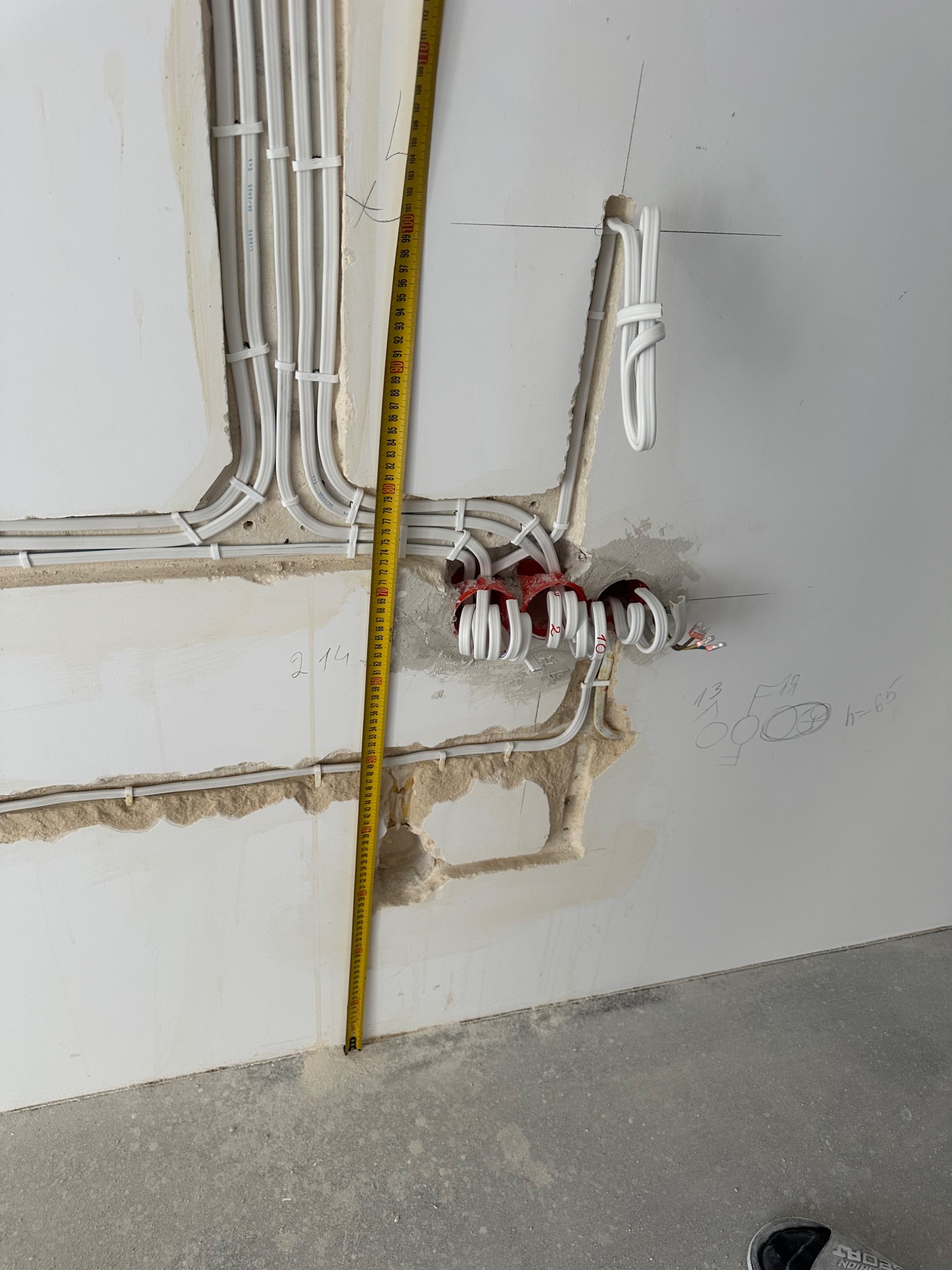 Białe przewody elektryczne ułożone w bruzdach w ścianie, zabezpieczone uchwytami. Widoczna miarka mierząca odległość od podłogi. Ściana częściowo otynkowana.