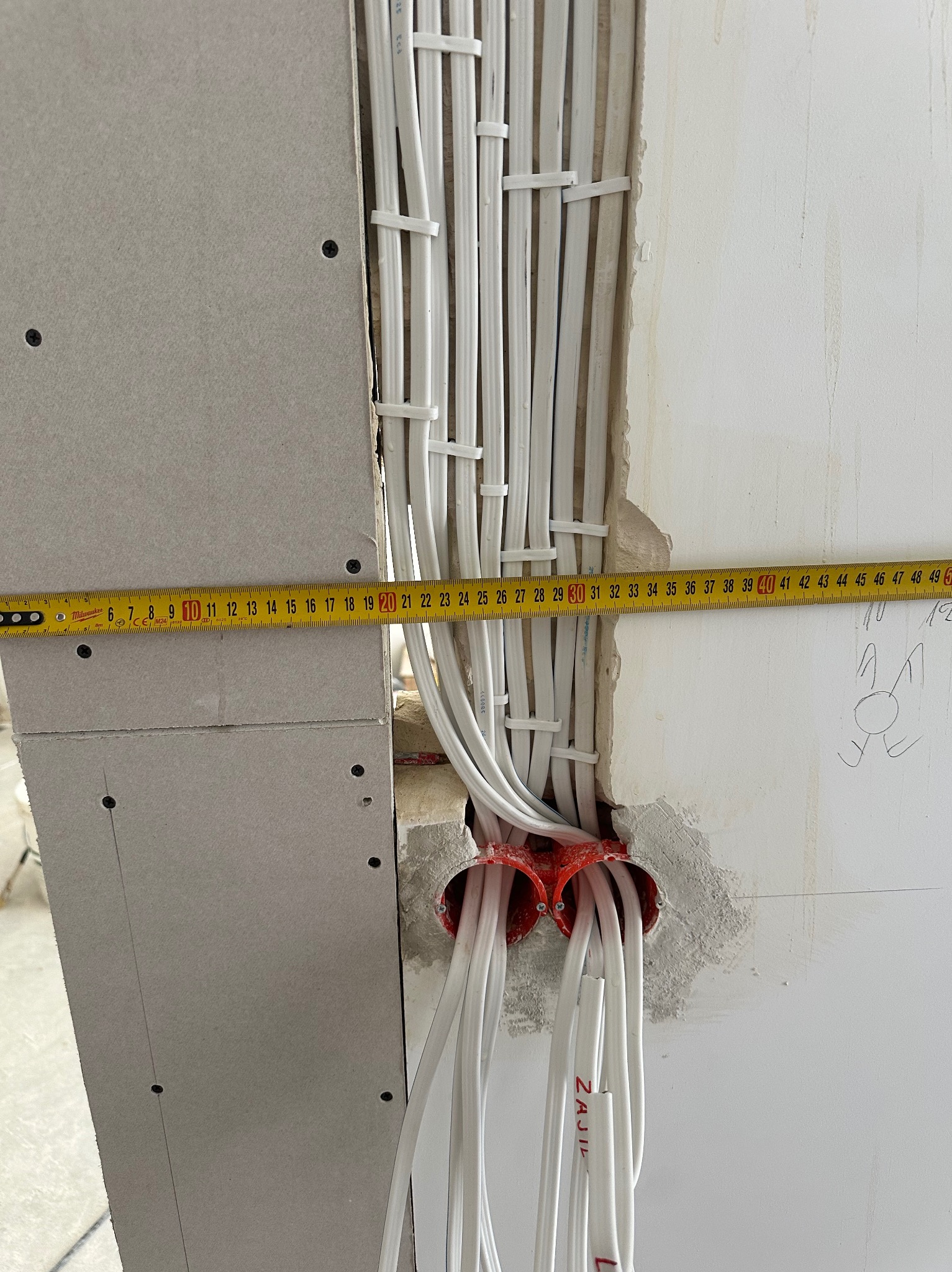 Białe kable elektryczne prowadzone w ścianie, zabezpieczone listwami i mierzone taśmą mierniczą. Widoczne puszki instalacyjne w kolorze czerwonym.