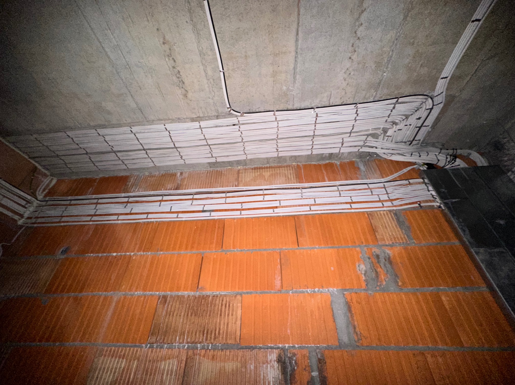 Białe kable elektryczne równolegle ułożone pod betonowym sufitem, nad czerwoną ścianą z cegieł. Instalacja w trakcie realizacji, widok z dołu.