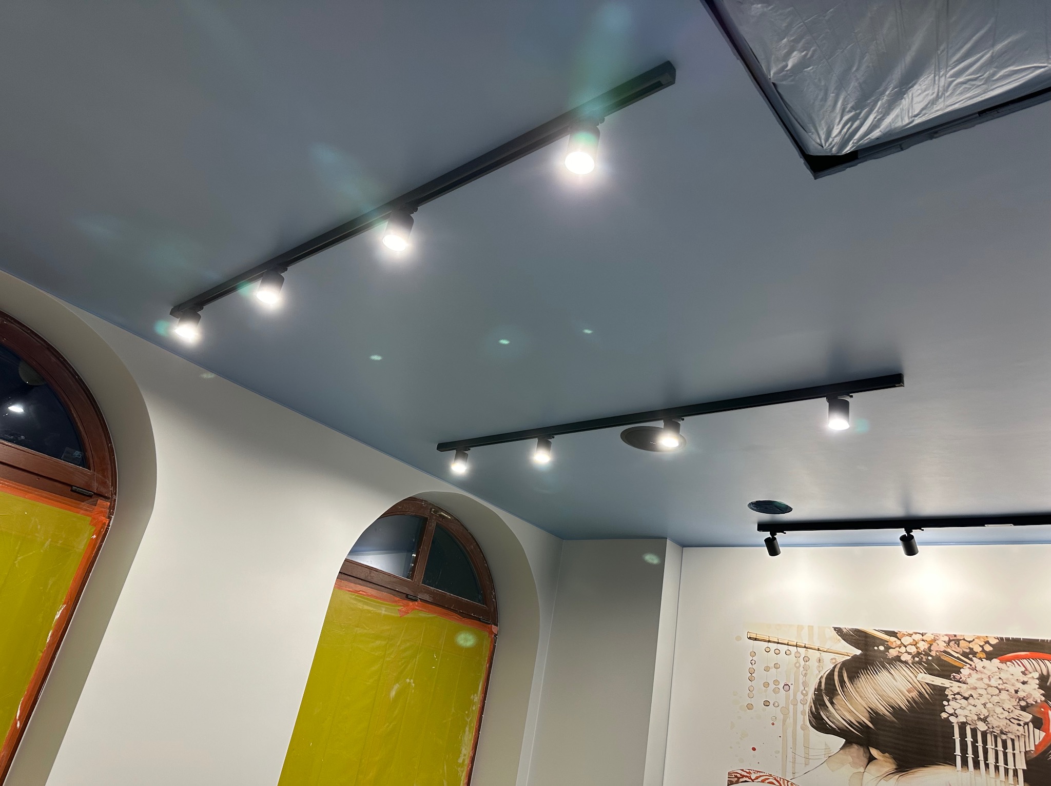 Nowoczesne oświetlenie szynowe z reflektorami w jasnym wnętrzu, okna zabezpieczone folią ochronną, obraz na ścianie. Niebieski sufit.