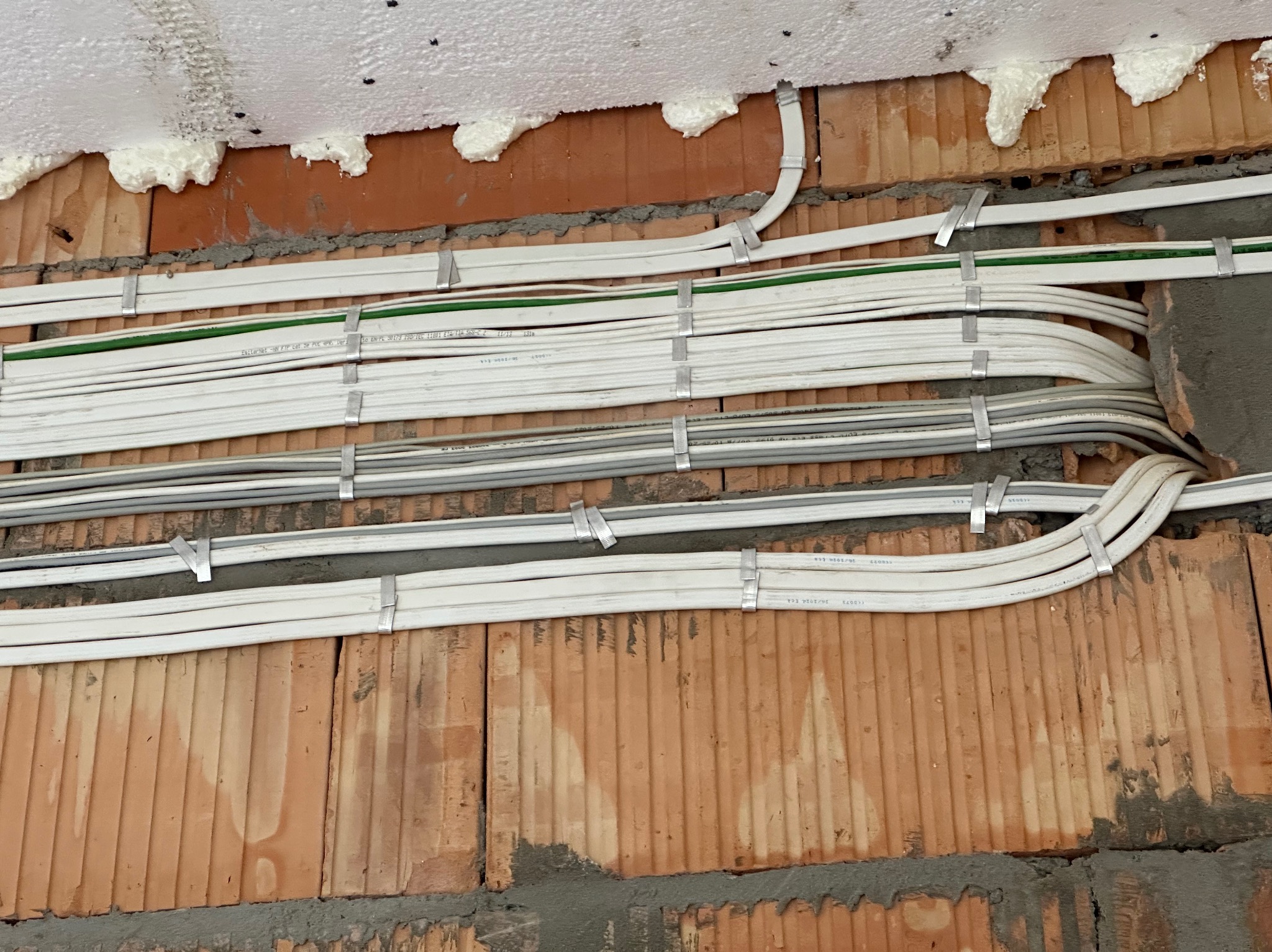 Kable elektryczne zamocowane klamrami na ścianie z cegły, przy suficie z pianką montażową. Instalacja w trakcie realizacji, widoczne oznaczenia na kablach.