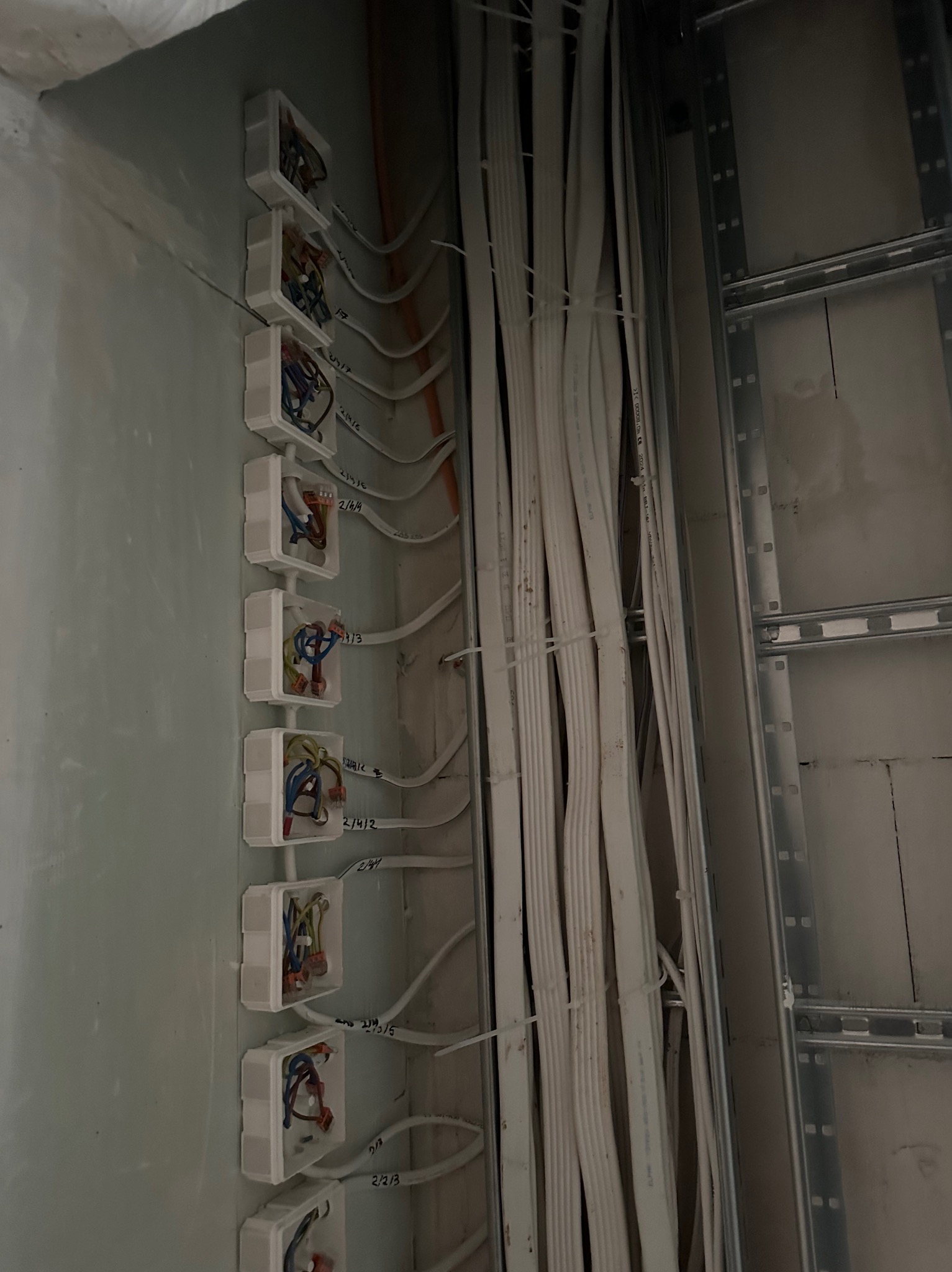 Rozprowadzenie przewodów elektrycznych w ścianie z puszkami instalacyjnymi. Widoczne kable oznaczone numerami, szyny montażowe. Prace elektryczne w surowym stanie.