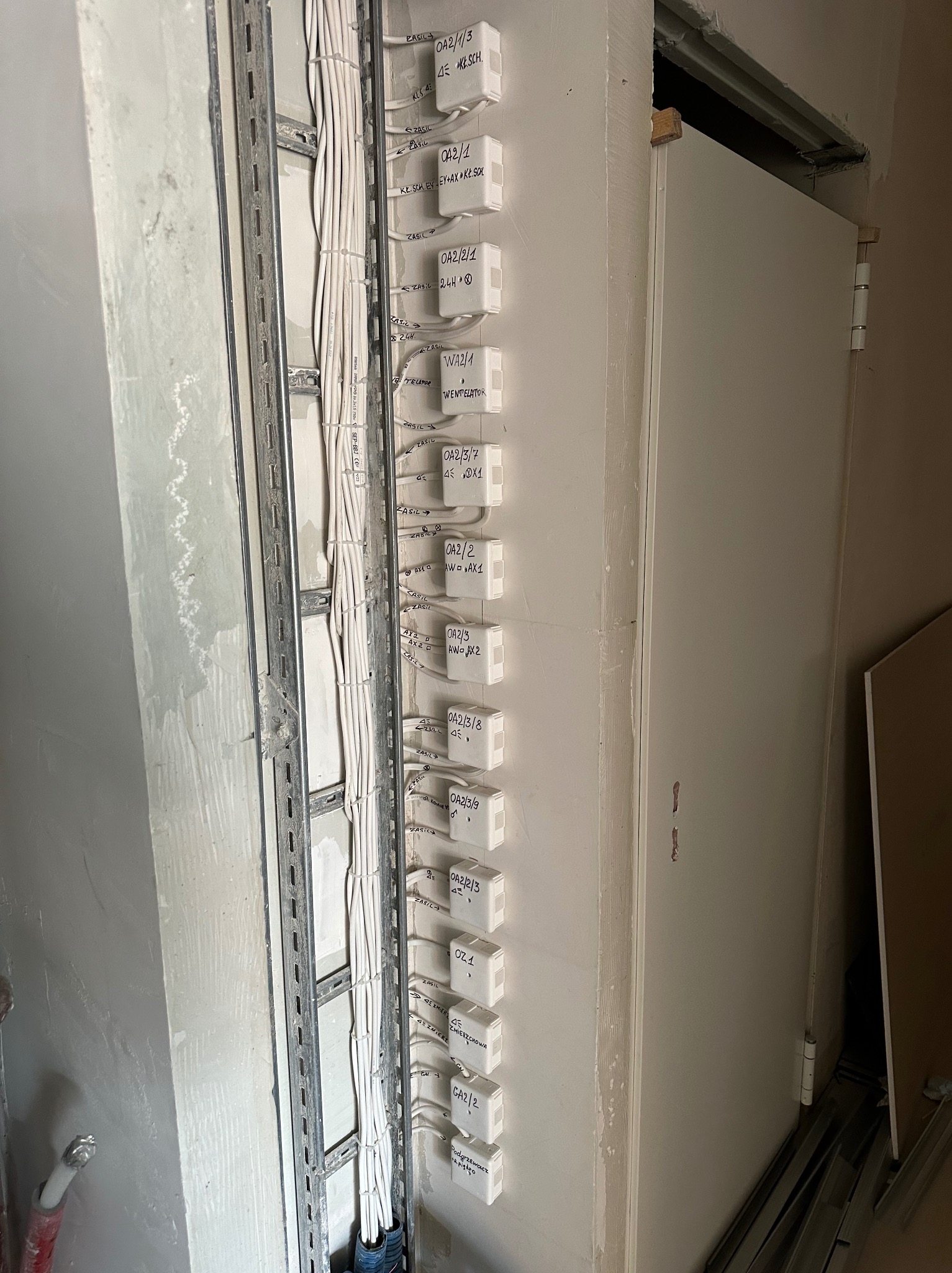 Rozprowadzenie kabli elektrycznych w korycie kablowym, z oznaczonymi puszkami instalacyjnymi na ścianie. Widoczne przewody i metalowa konstrukcja mocująca.