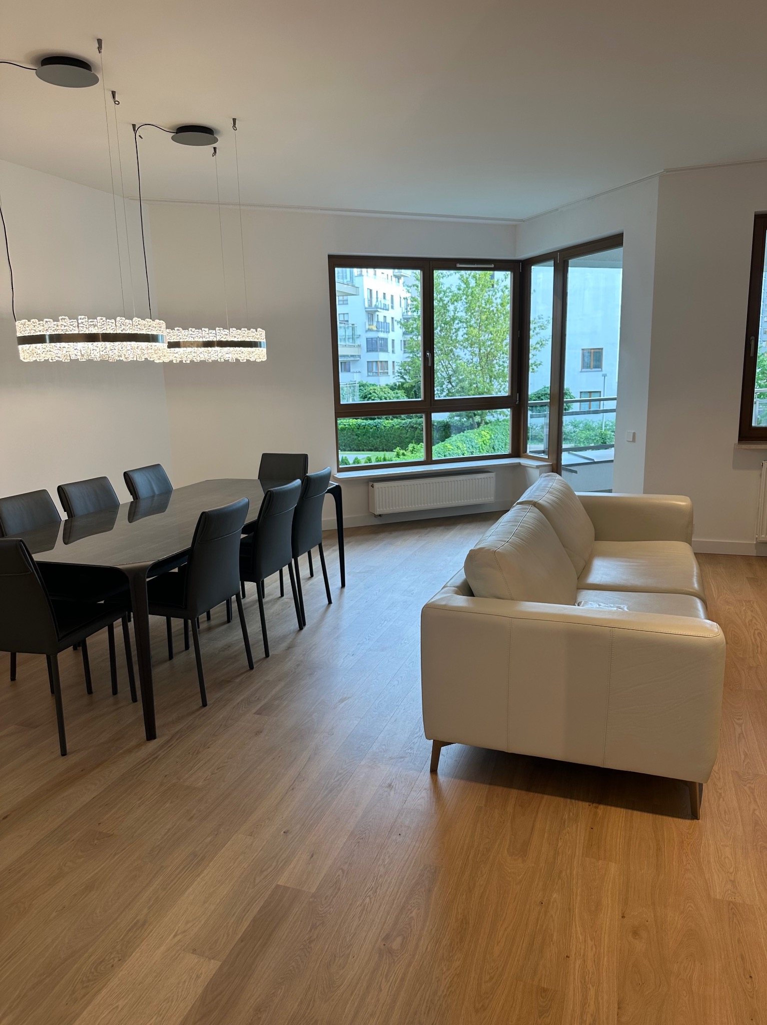 Nowoczesne wnętrze: stół z krzesłami i skórzana sofa w przestronnym salonie z dużymi oknami i parkietem. Nad stołem designerskie, kryształowe lampy.