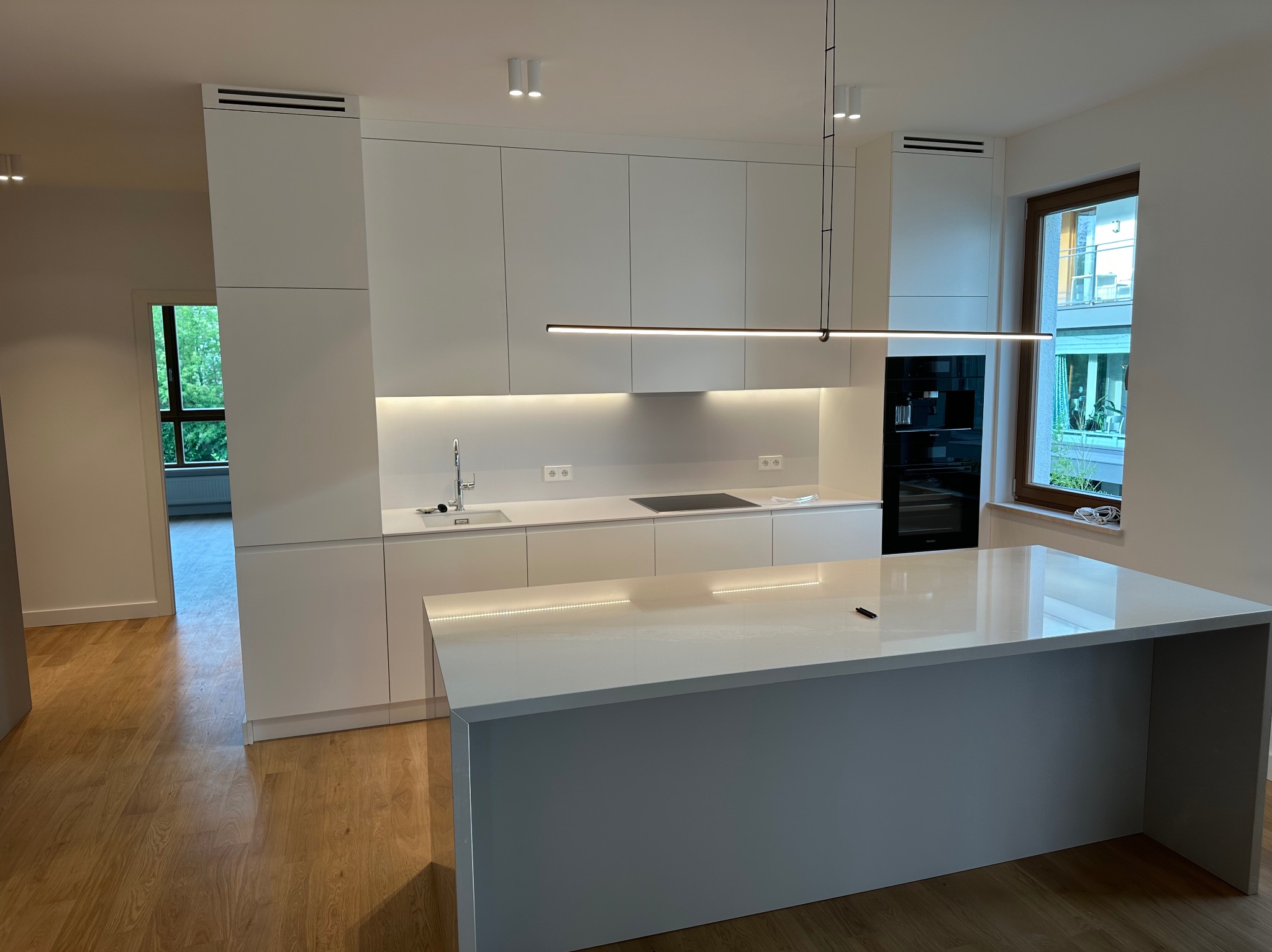 Minimalistyczna, biała kuchnia z wyspą, oświetleniem LED podszafkowym i nowoczesną lampą wiszącą. Widok na okno i fragment salonu.