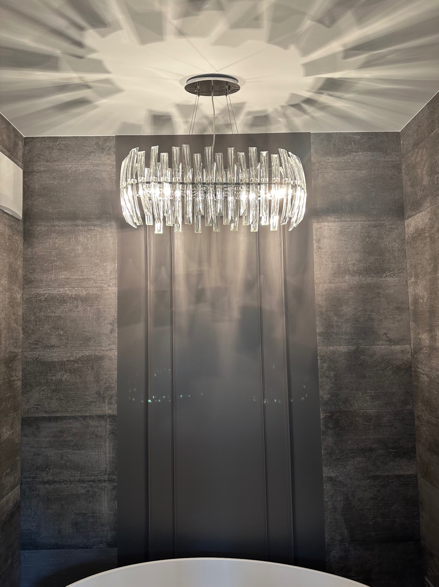 Kryształowy żyrandol w nowoczesnej łazience z szarymi płytkami. Światło tworzy dekoracyjny efekt na suficie. Widoczny fragment wanny.