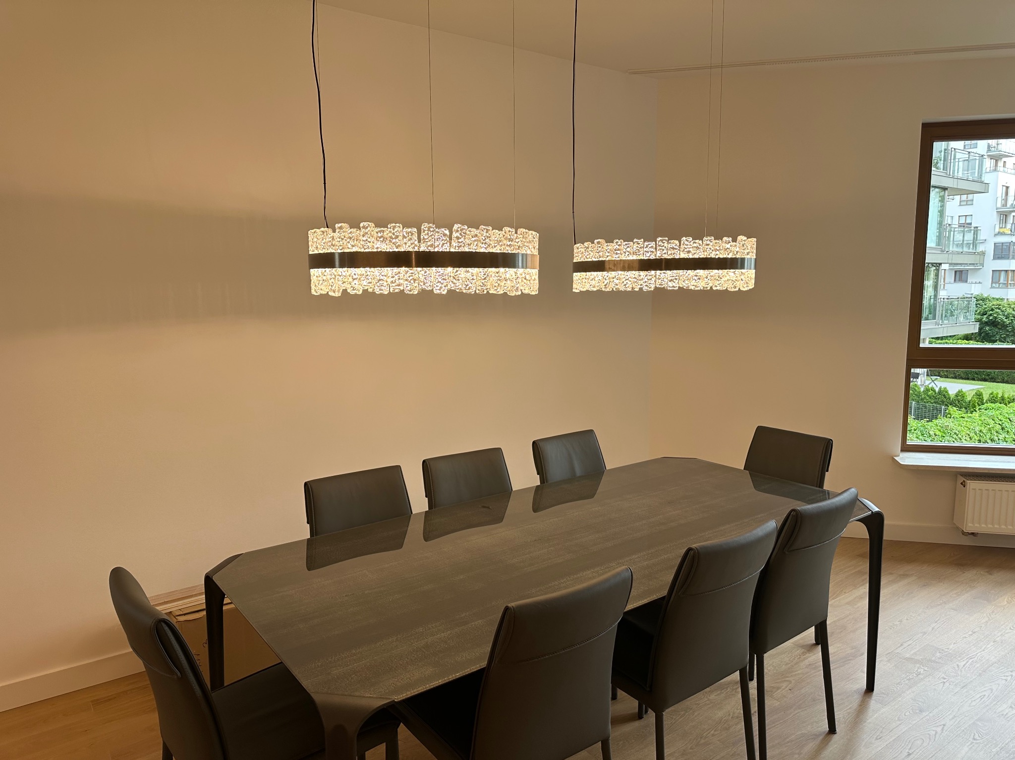 Dwa nowoczesne, okrągłe żyrandole z kryształkami nad długim stołem z krzesłami w jasnym wnętrzu. Widok na zieleń za oknem.