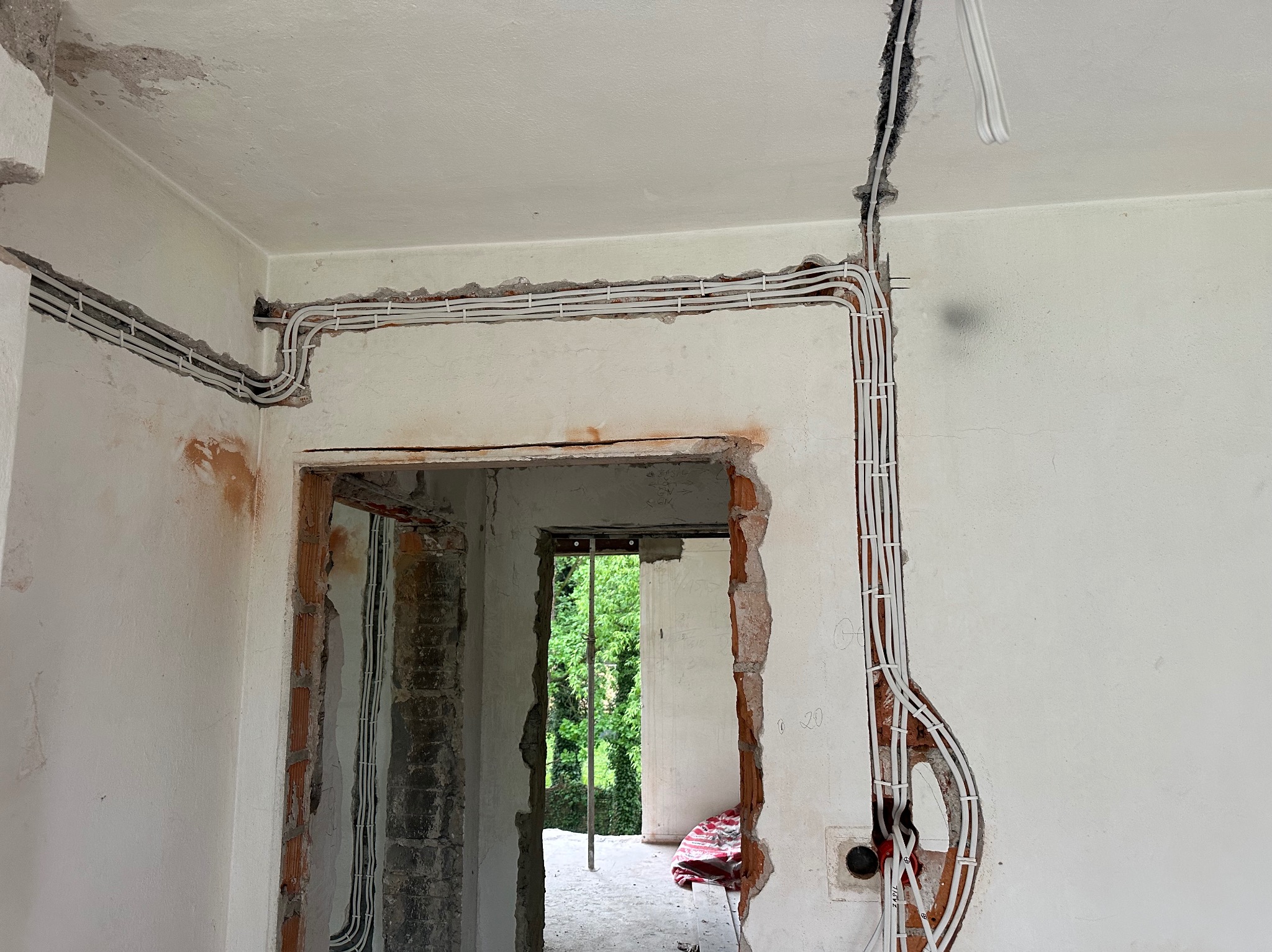 Białe kable elektryczne ułożone w bruzdach ścian w trakcie remontu. Widoczne fragmenty cegieł, instalacja w peszlu i widok na zieleń przez otwór drzwiowy.
