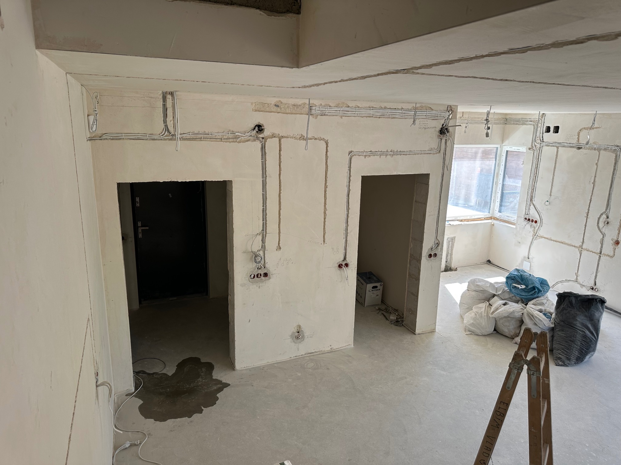 Wnętrze w trakcie remontu: widoczne ściany z instalacją elektryczną w peszlach, otwory na gniazdka, surowy betonowy sufit, worki z gruzem i drabina w pomieszczeniu.
