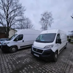 Dwa białe busy dostawcze Fiat Ducato zaparkowane na brukowanym placu w pochmurny dzień, widoczne tablice rejestracyjne EP 87983.