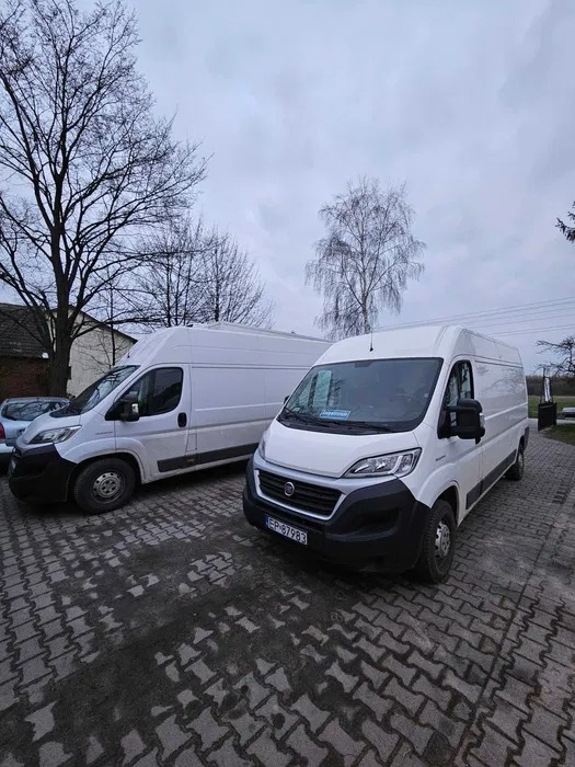 Dwa białe busy dostawcze Fiat Ducato zaparkowane na brukowanym placu w pochmurny dzień, widoczne tablice rejestracyjne EP 87983.